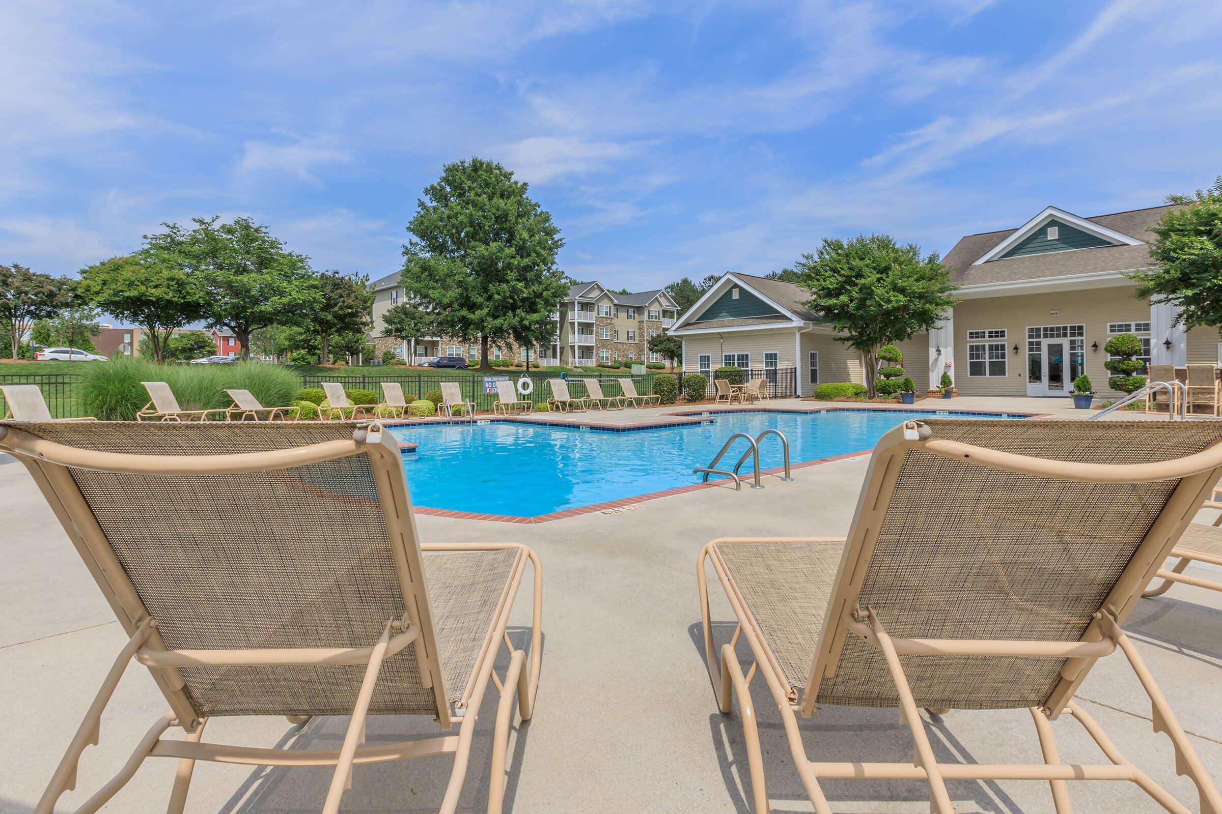 A serene pool area featuring a sparkling blue swimming pool surrounded by lounge chairs. Lush greenery and trees provide a natural backdrop, while a well-maintained building with large windows and a covered entrance is visible in the background under a clear blue sky.