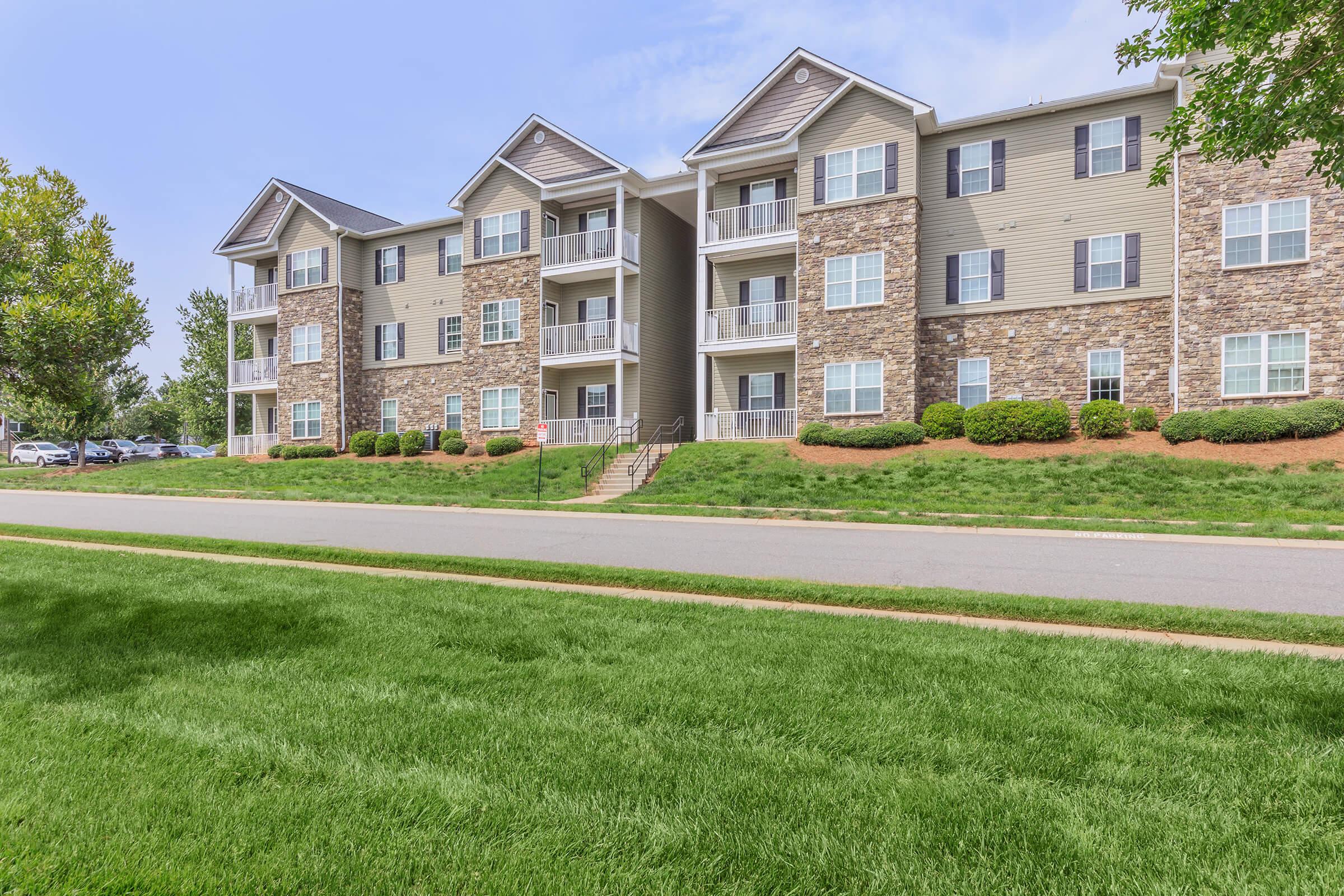 A row of modern, multi-story apartment buildings with stone accents, surrounded by well-manicured green lawns and shrubs. The buildings feature balconies and many windows, under a clear blue sky. A paved road runs alongside the property, with parking spaces visible in the background.