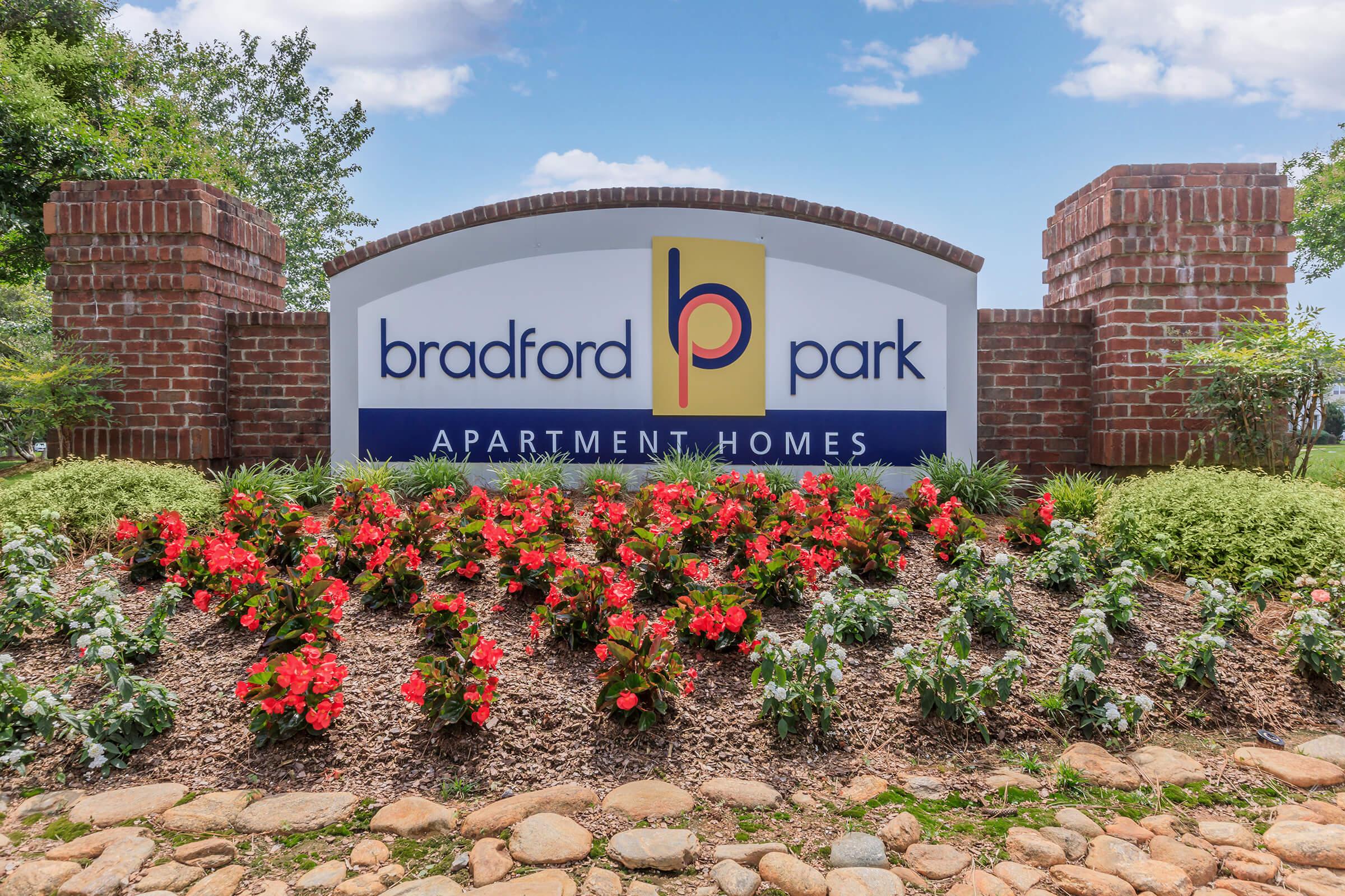 Sign for Bradford Park Apartment Homes, featuring a large logo with the letter "P" and surrounded by landscaped flower beds with colorful blooms. The sign is flanked by brick columns, set against a clear blue sky and greenery in the background.
