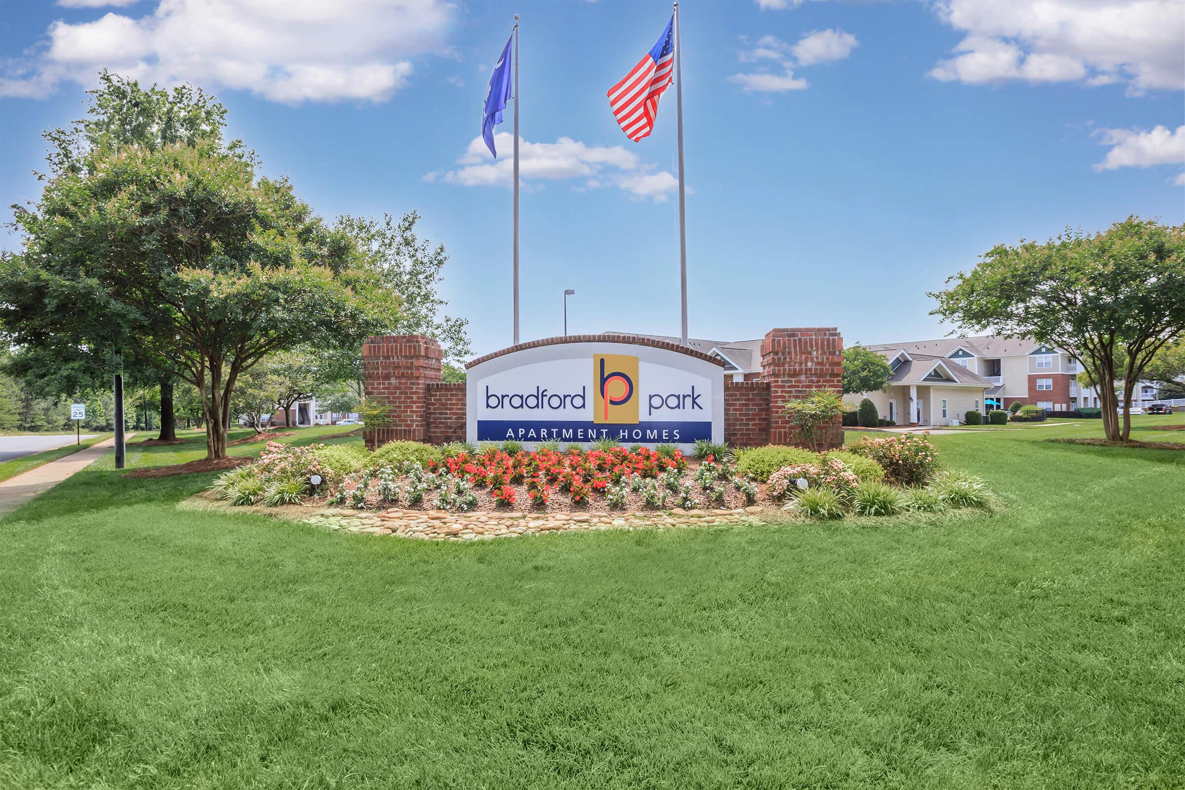 A welcoming sign for Bradford Park Apartment Homes, surrounded by colorful flowers and lush greenery, with two flags flying in the background under a bright blue sky.