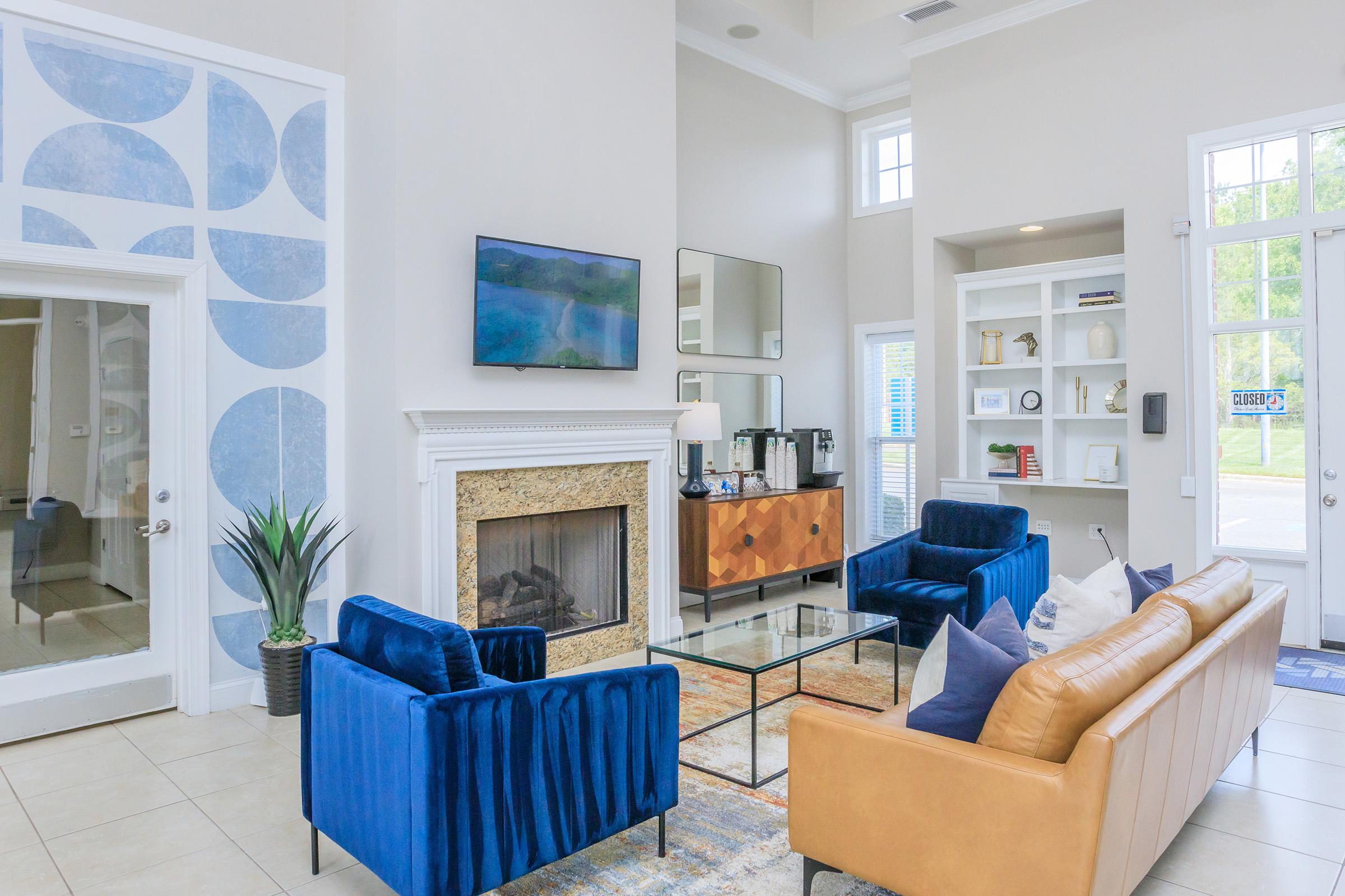 A modern living area featuring a cozy arrangement of blue and brown sofas, a glass coffee table, and a stone fireplace. There is a flat-screen TV mounted on the wall and a decorative shelf with books and decor. Large windows allow natural light into the space, creating a welcoming atmosphere.