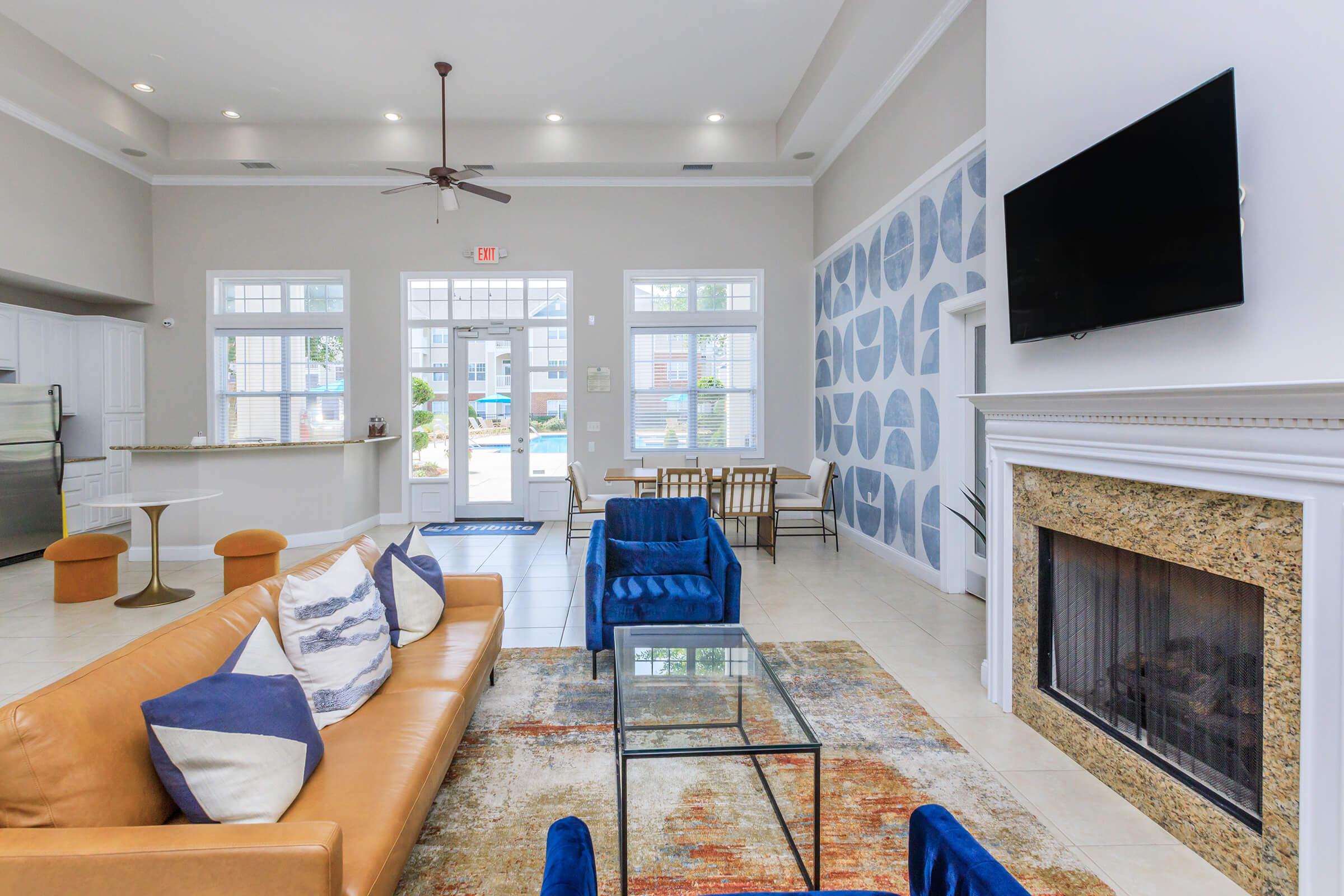 A spacious modern living area featuring a tan leather sofa with decorative pillows, a glass coffee table, and a cozy fireplace. The room is brightly lit with large windows, and there are blue accent chairs. In the background, a dining table and a kitchen area are visible, creating a welcoming atmosphere.