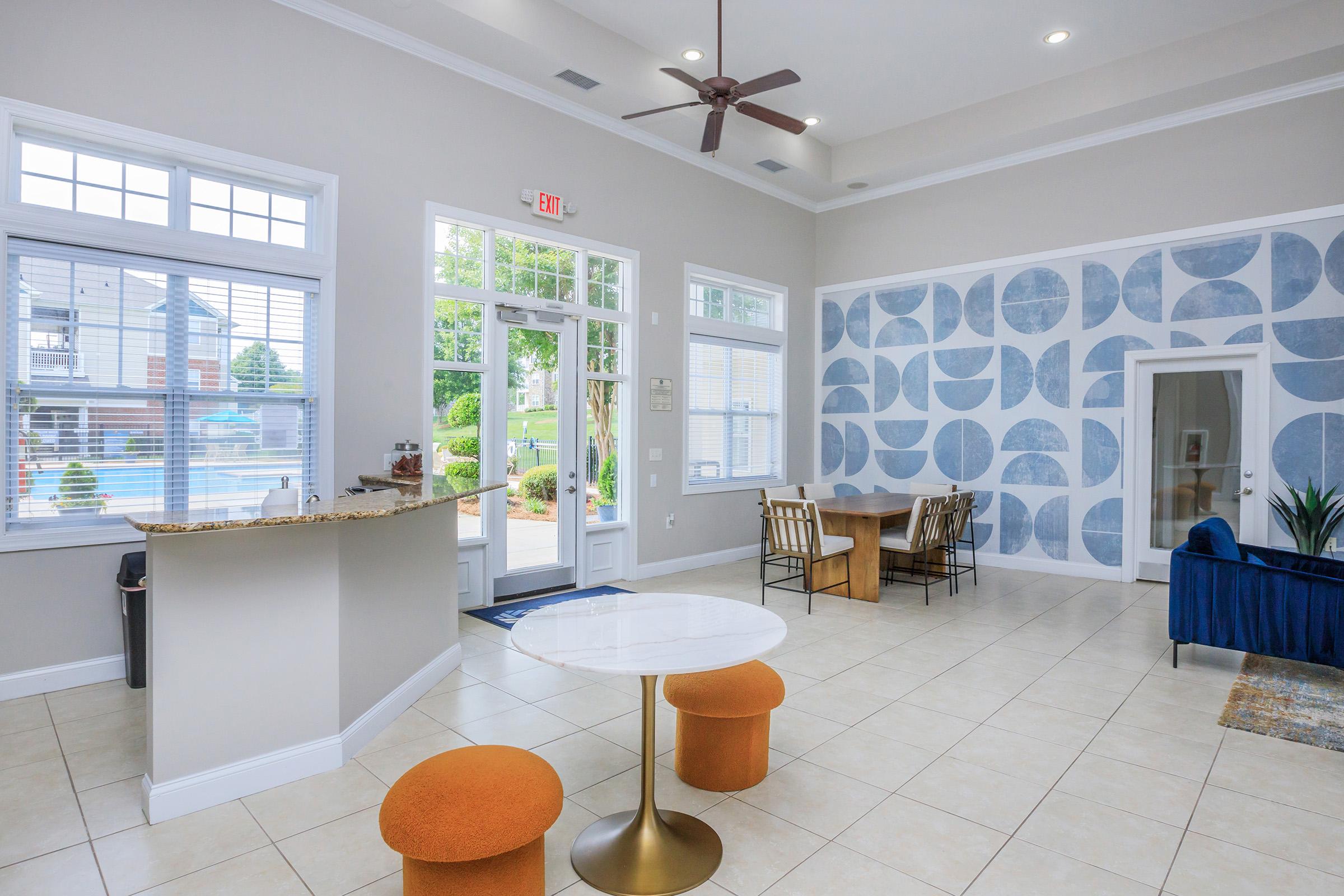A modern lobby area featuring a marble table with round orange stools, a wooden dining table, and a blue couch. Large windows provide natural light, revealing a pool outside. The walls are decorated with a geometric blue pattern. A reception area with a granite counter is visible.