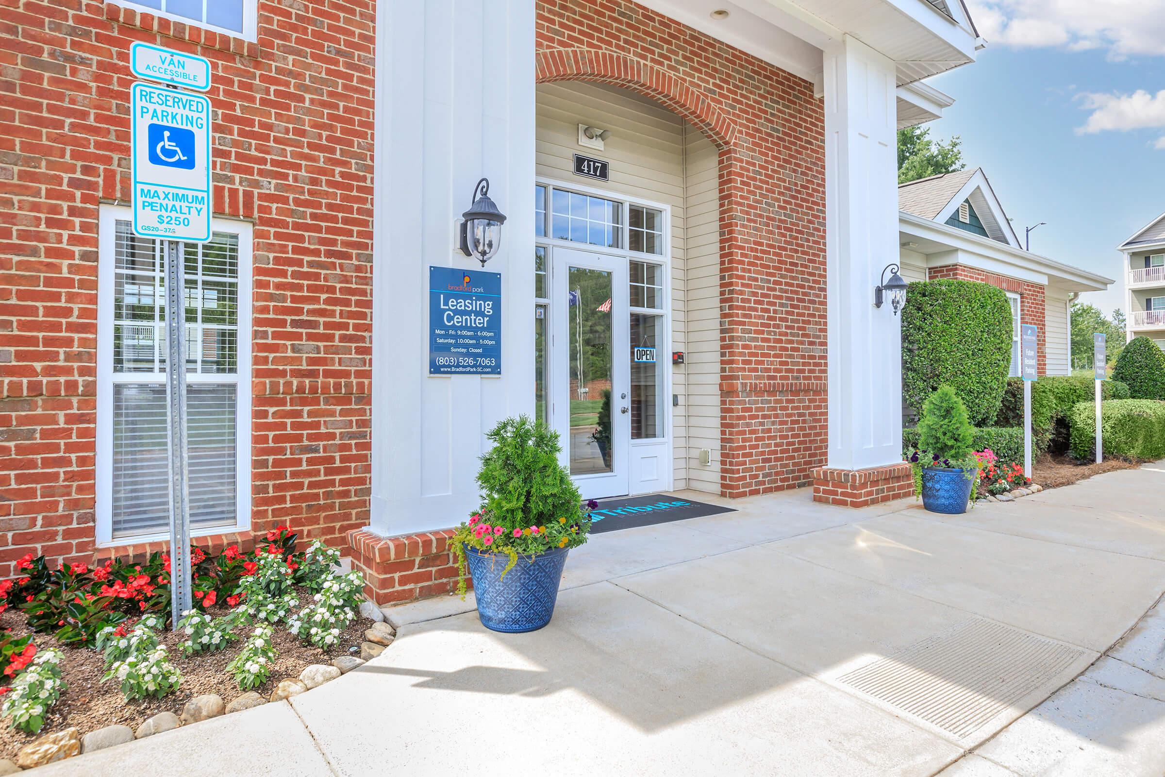 Entrance to a Leasing Center featuring a brick exterior, large columns, and a colorful sign. There are decorative planters with flowers on either side of the entrance and a sidewalk leading up to the door. Parking signs are visible, including one for reserved parking.