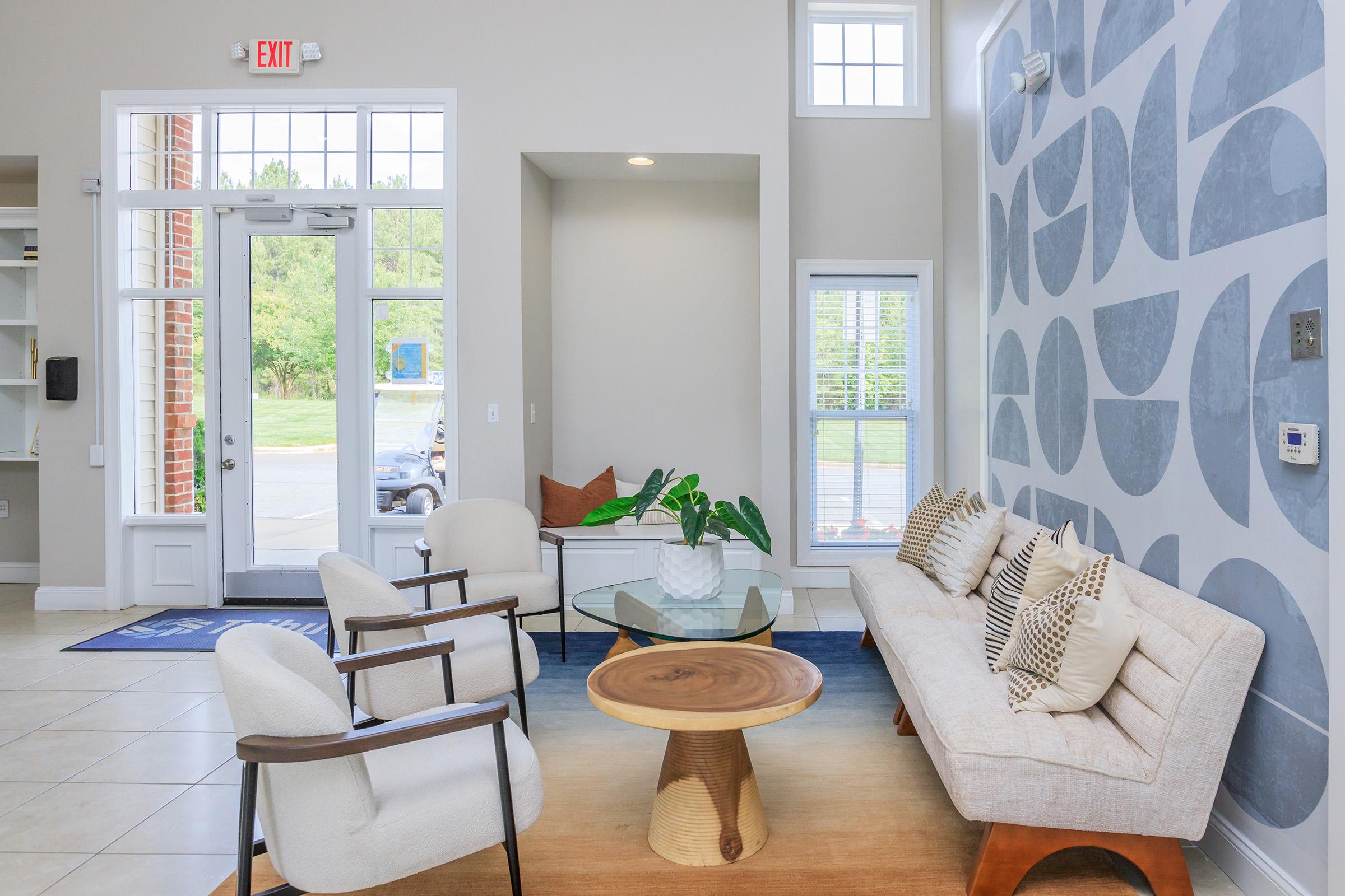 Modern lobby area featuring a light color palette. A stylish beige sofa and two wooden chairs surround a round wooden coffee table. There’s a large window letting in natural light and a decorative plant on the table. The wall has a contemporary design with circular patterns in soft colors.