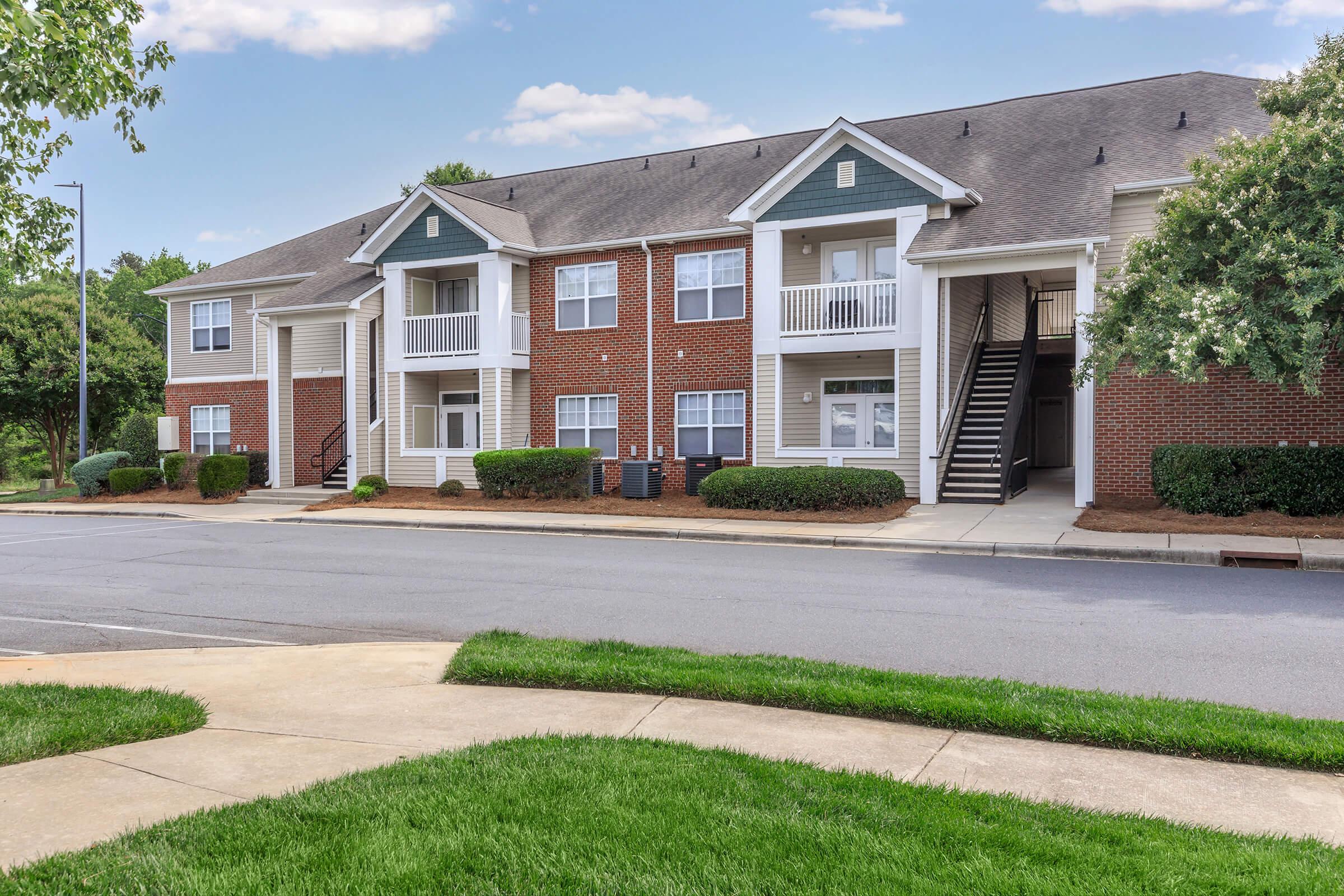 A two-story brick apartment building with white balconies and green shutters, surrounded by well-manicured lawns and shrubs. The building features a sidewalk in front and a parking area nearby. Clear blue skies are visible above, indicating a sunny day.
