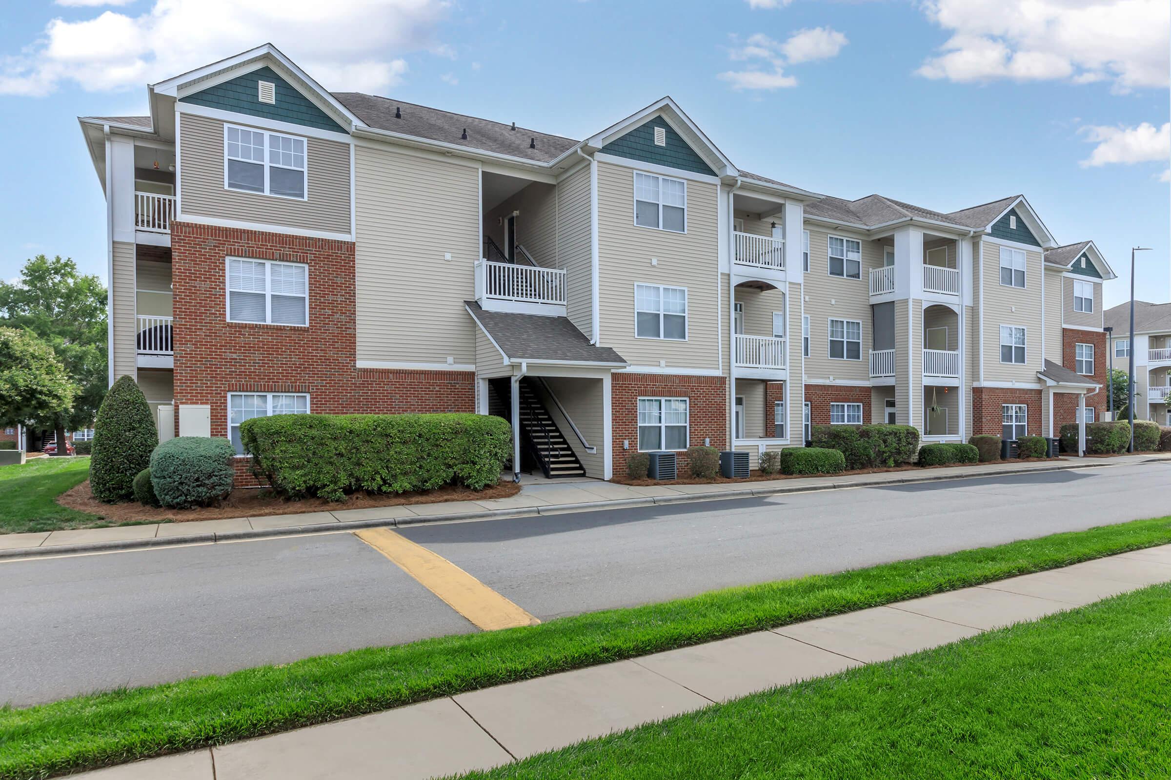 A three-story residential apartment building featuring brick and siding exterior, with balconies on the second and third floors. Well-maintained landscaping includes trimmed hedges and a walkway alongside the building, set against a clear blue sky.