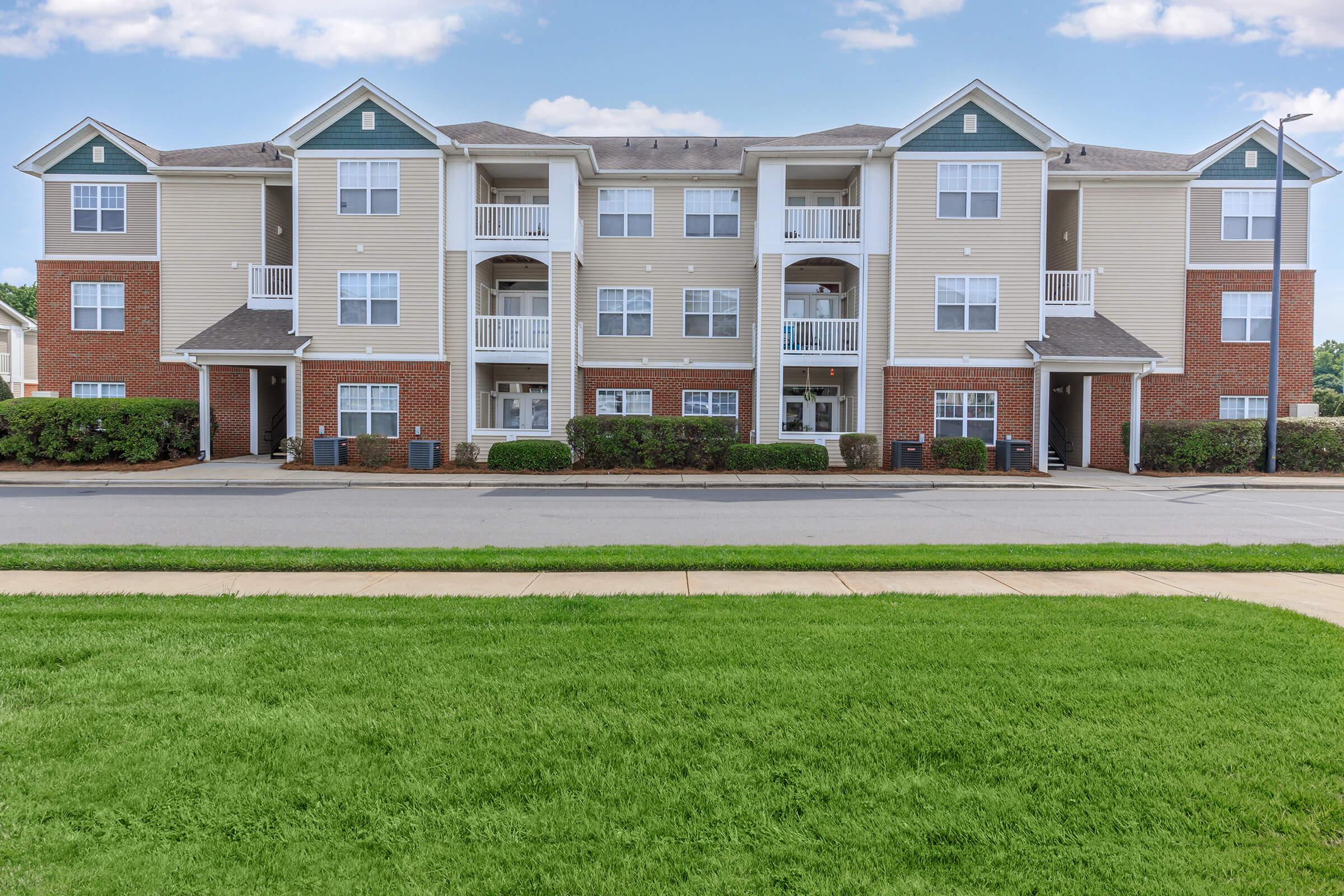 A three-story residential apartment building featuring a combination of brick and siding exteriors. Balconies are visible on the upper floors, and the ground is landscaped with neat grass and shrubs. The building is organized with a symmetrical layout and multiple entrances.