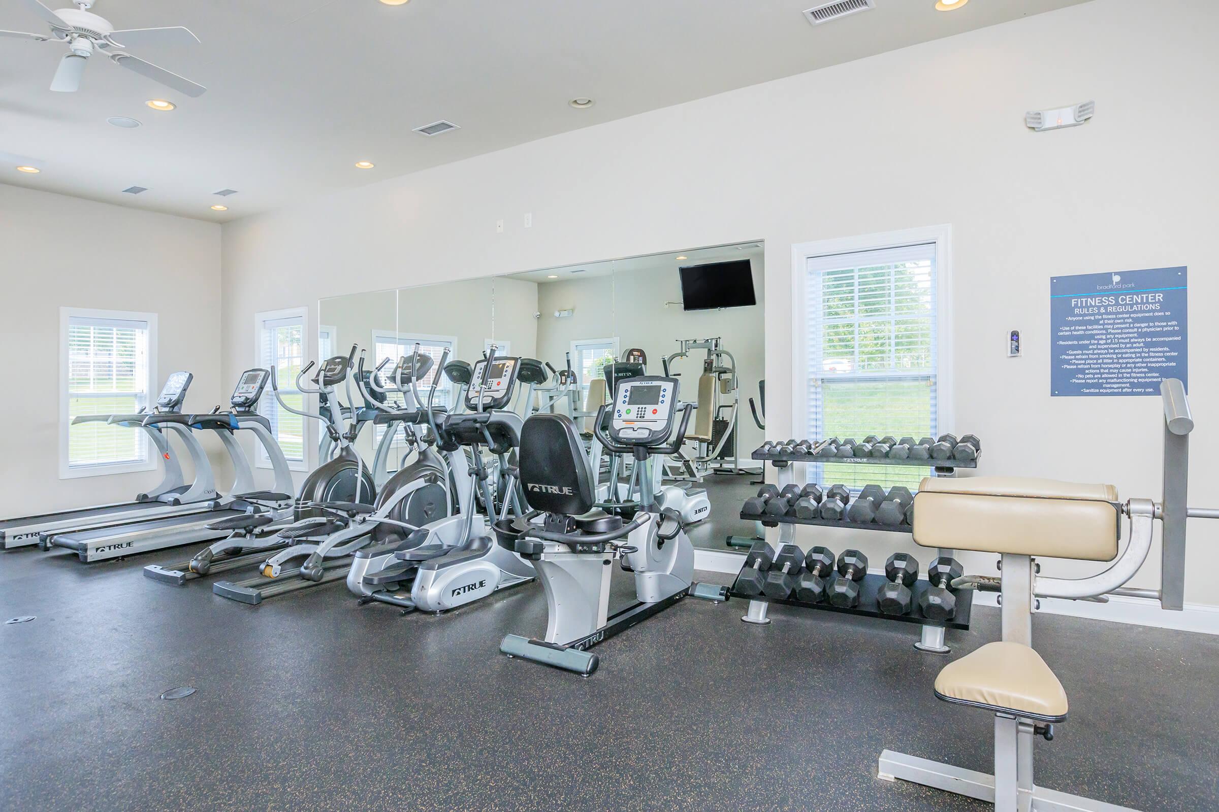 Modern fitness center featuring cardio machines, including treadmills and elliptical trainers, along with free weights. Bright, airy space with natural light, equipped with a wall-mounted TV and mirrors reflecting the workout area. Motivational signage is visible in the background.