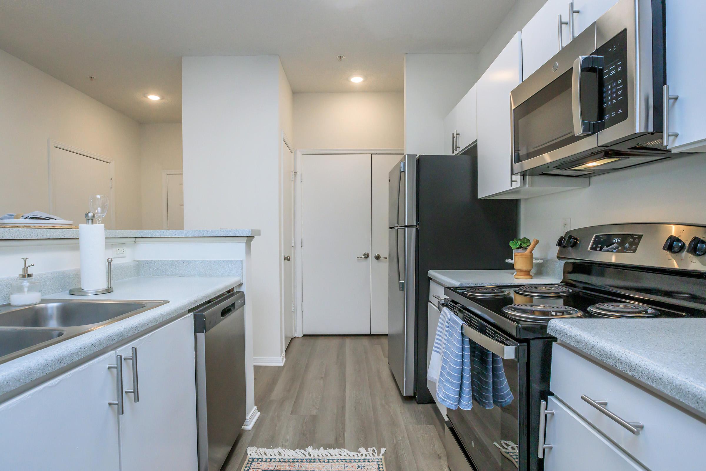 A modern kitchen featuring white cabinets, stainless steel appliances, a countertop with a double sink, and a microwave. The space has a sleek design with a black refrigerator, an oven, and a stove. Soft lighting illuminates the area, and a door leads to another room in the background.