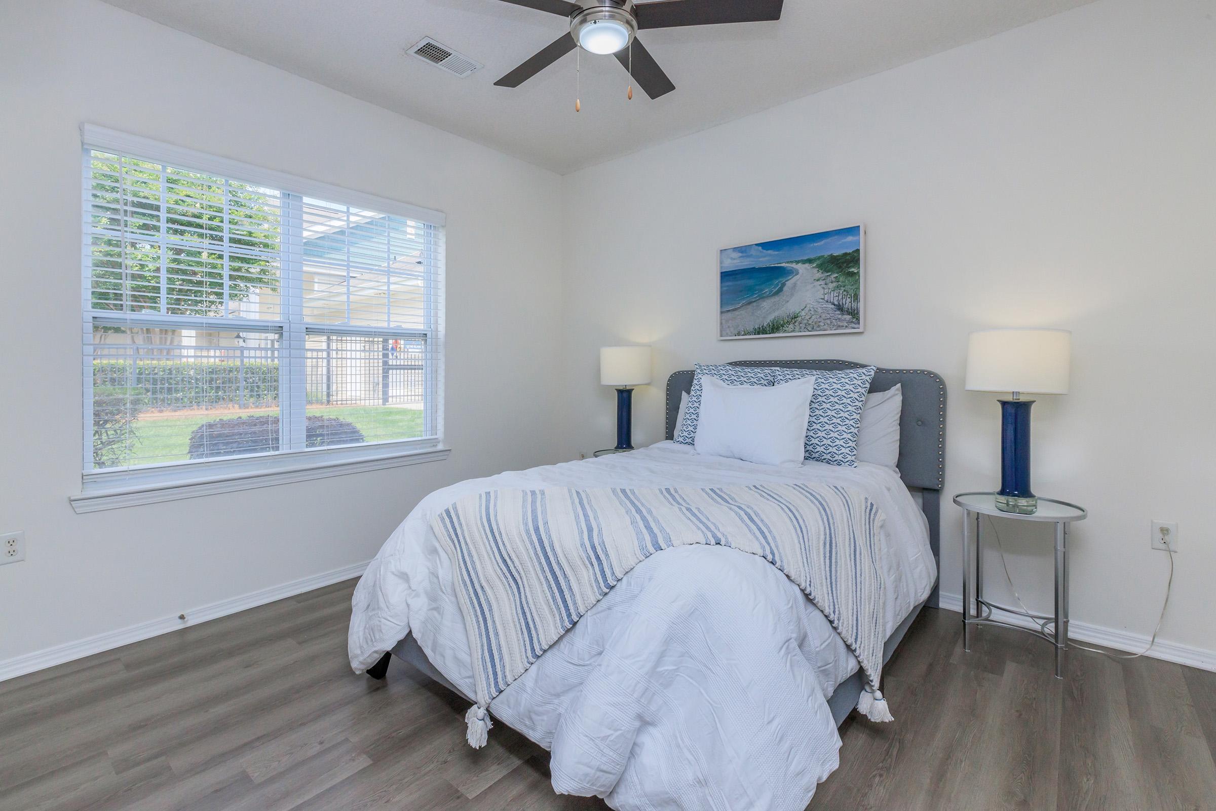 A cozy bedroom featuring a neatly made bed with white and blue bedding, two bedside lamps on either side, and a window letting in natural light. A beach-themed painting hangs above the bed, and the room has light wood flooring and a ceiling fan, creating a bright and inviting atmosphere.