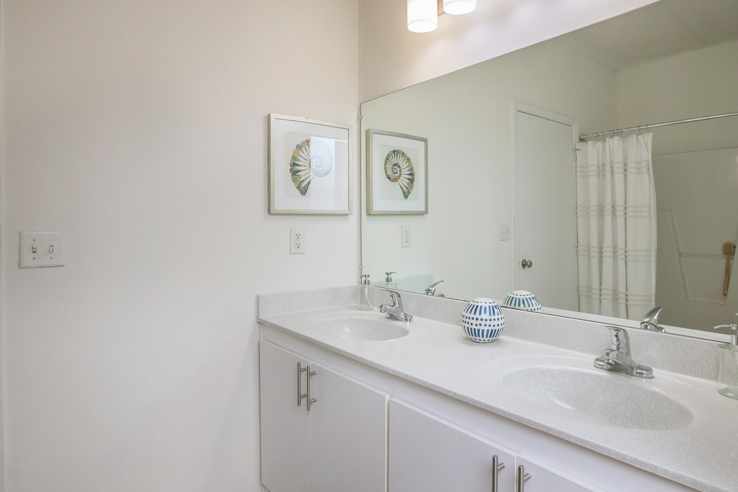 A bright and modern bathroom featuring a double sink vanity with sleek silver faucets. Above the sinks, two decorative framed artworks are displayed. A large mirror reflects the space, and a light fixture with multiple bulbs illuminates the area. A shower curtain is visible in the background.