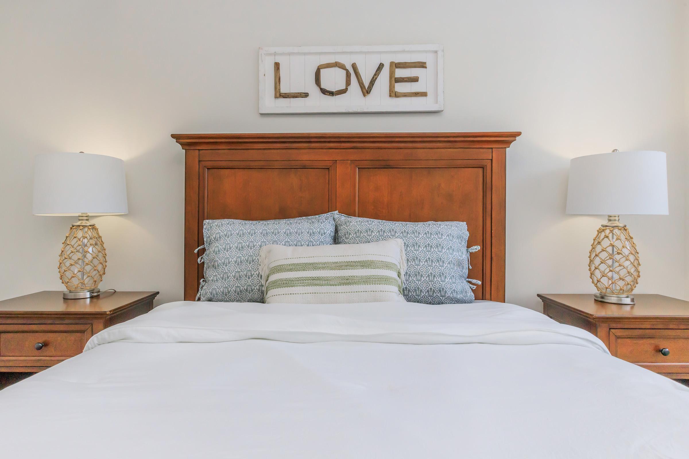 A cozy bedroom featuring a wooden headboard with a "LOVE" sign above it. The bed is neatly made with white linens and decorative pillows in blue and green tones. On either side of the bed, there are wooden nightstands with stylish lamps that have a woven design. The walls are painted a soft color.