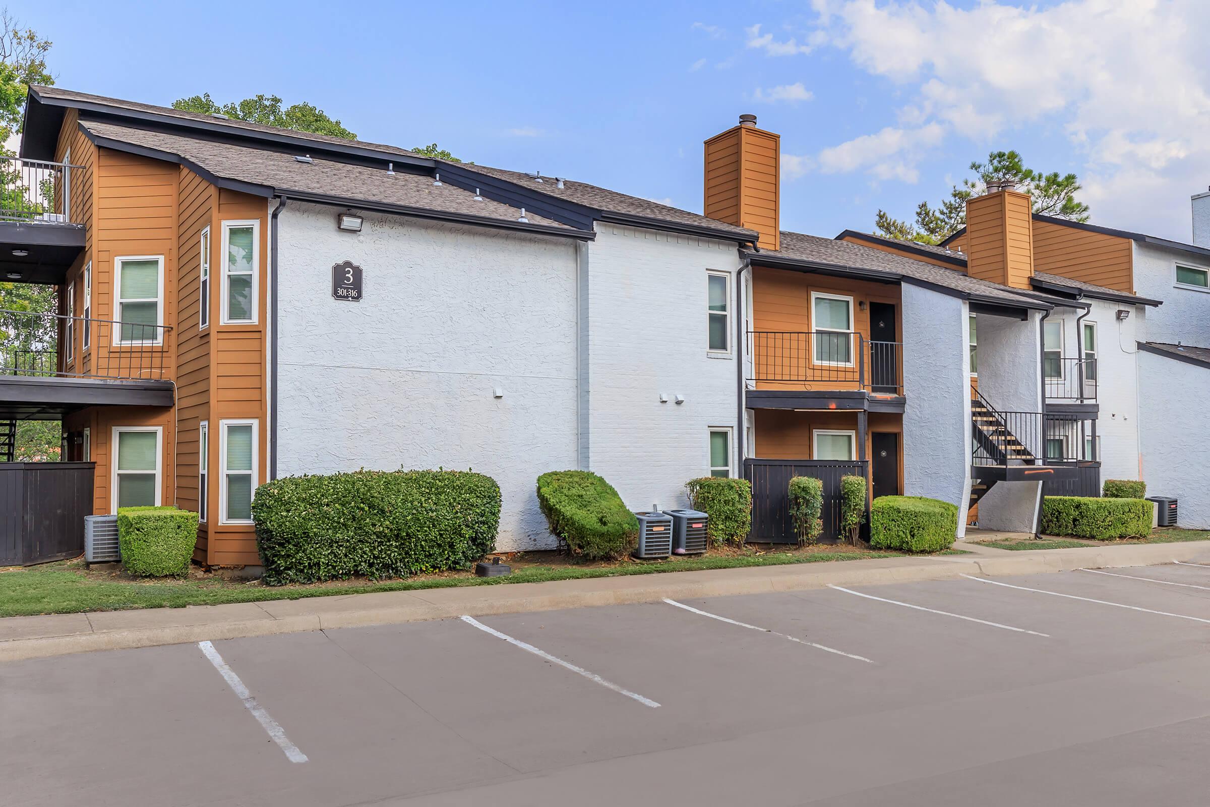Exterior view of a multi-unit apartment building featuring a combination of orange and white siding. The building has multiple balconies, air conditioning units, and a landscaped area with shrubs. A parking lot is visible in front, with several empty parking spaces.