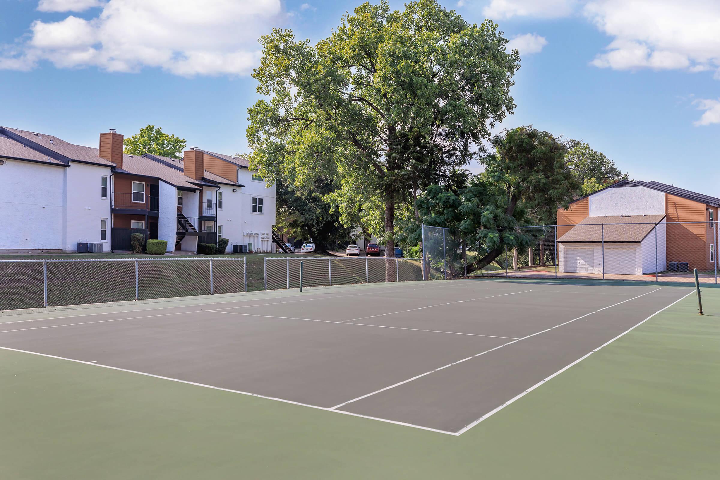 A view of a tennis court surrounded by residential buildings and trees, under a clear blue sky. The court features a gray playing surface with boundary lines, and is situated next to a chain-link fence. In the background, several apartments are visible alongside parked cars.