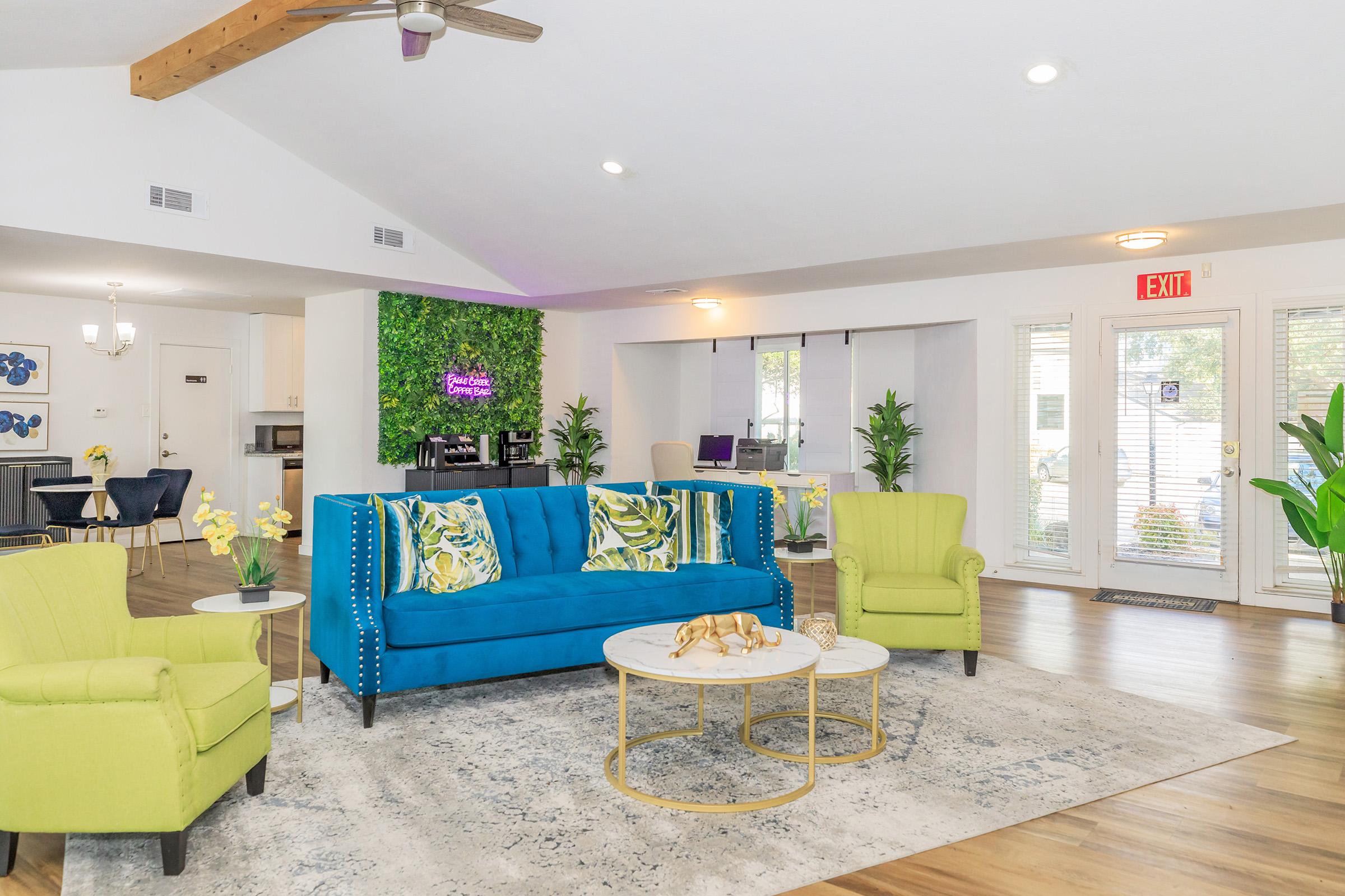 A modern living space featuring a bright blue sofa with patterned cushions, two vibrant green armchairs, and a round coffee table. The room has large windows letting in natural light, indoor plants, and a green accent wall with decorative elements. A dining area is visible in the background.