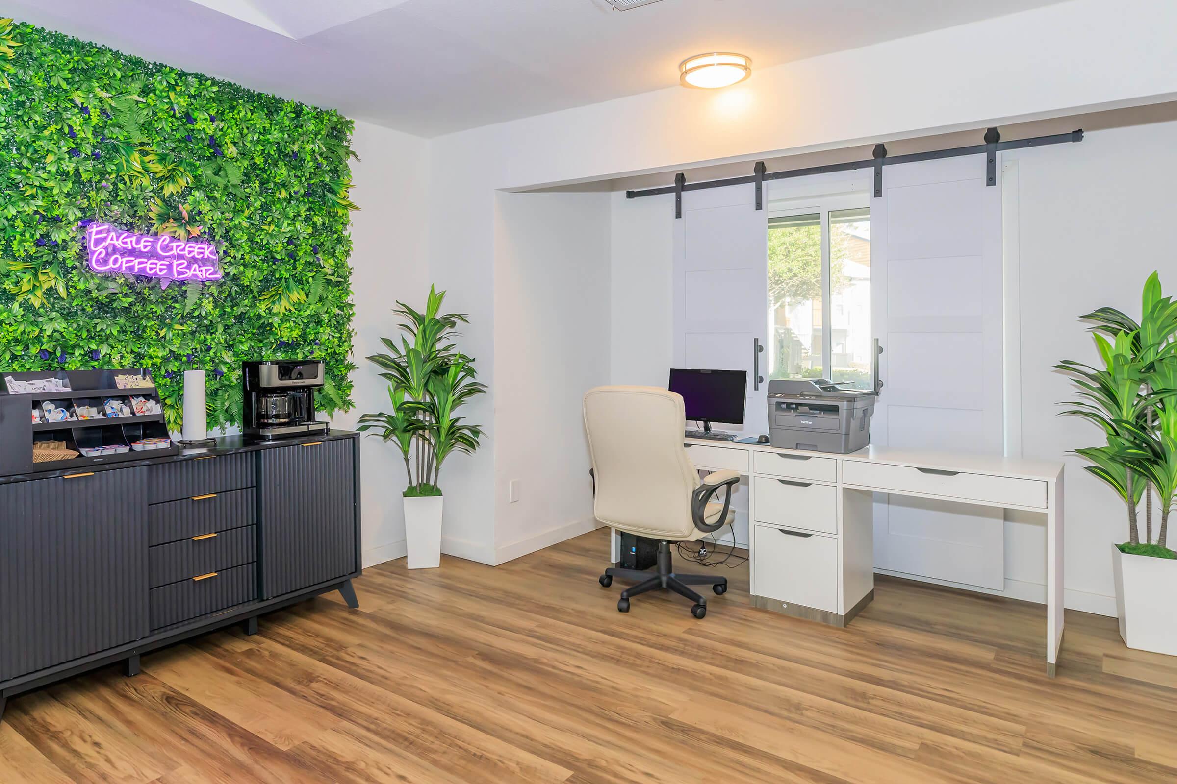 A modern office space featuring a vibrant greenery wall with a neon sign that says "Eagle Creek Coffee Bar." There’s a sleek black cabinet, a coffee machine, an office chair, and a desk with a printer, set against light-colored walls and a window allowing natural light.