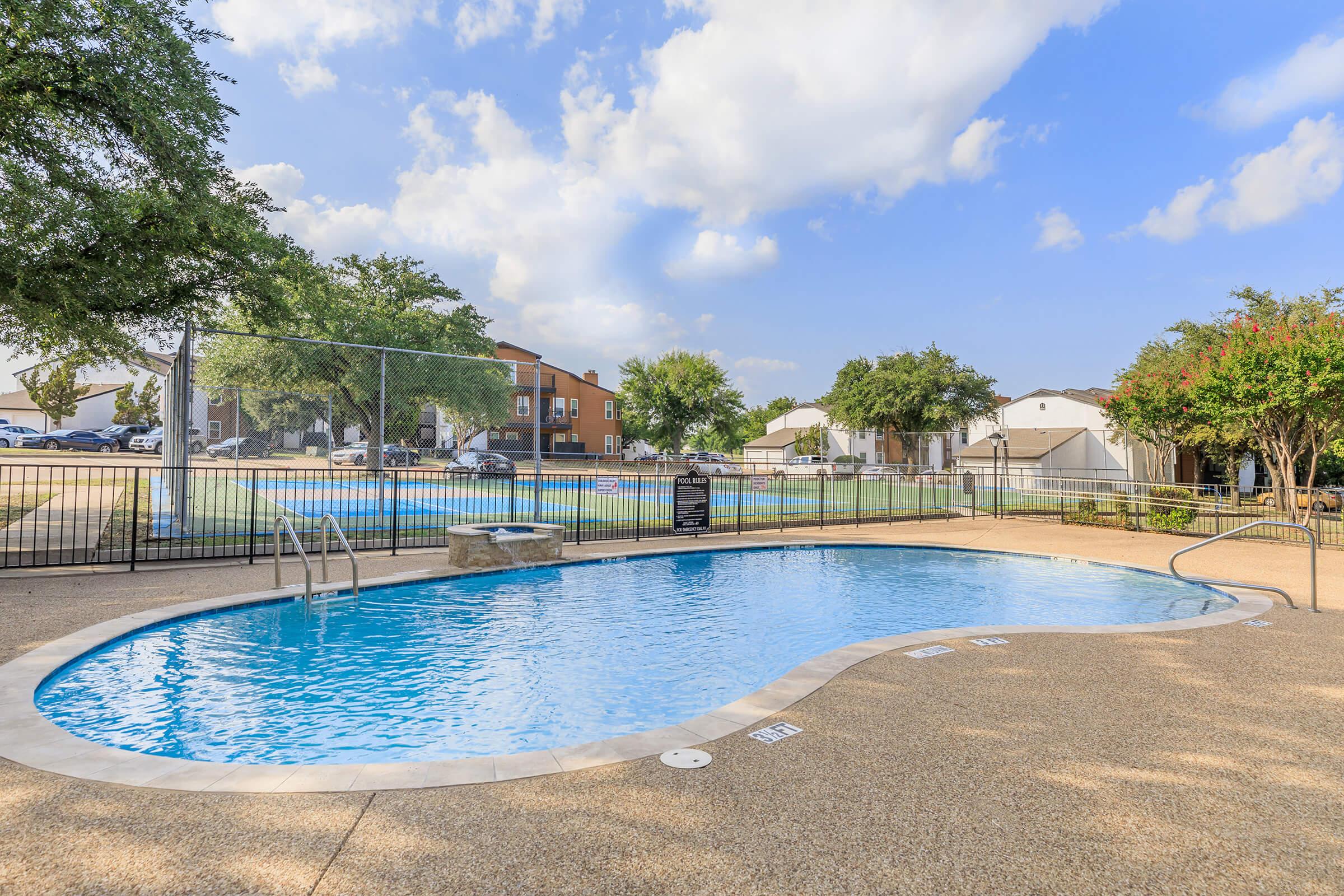 A sparkling swimming pool surrounded by a fenced area, featuring a textured concrete deck. The pool has a gentle curve and is inviting with clear blue water. In the background, there are residential buildings and lush green trees under a partly cloudy sky.