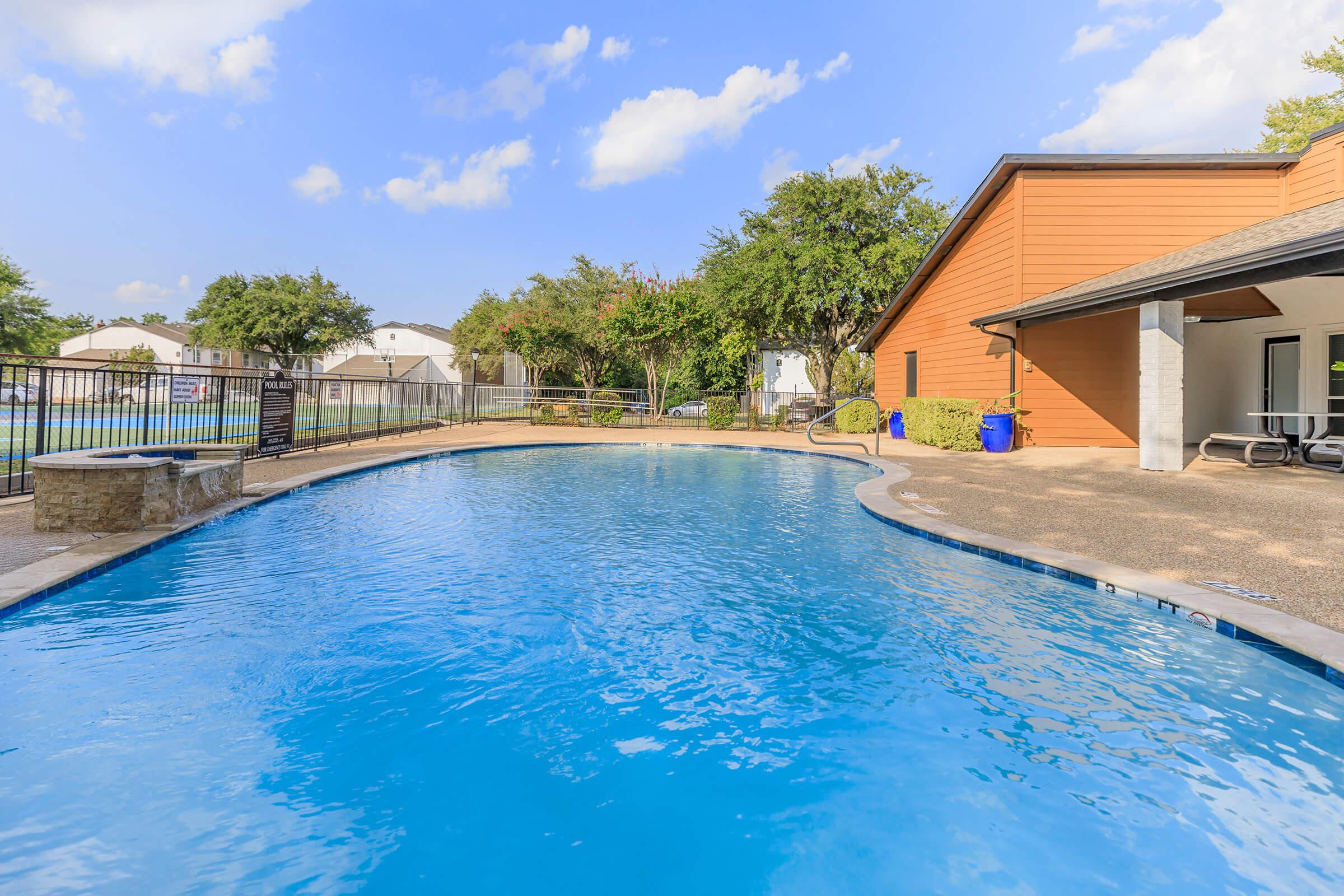 A clear blue swimming pool surrounded by a stone edge, with a wooden building on the right. Lush green trees line the background, and the sky is partly cloudy. A few lounge chairs are positioned nearby, creating a relaxing outdoor atmosphere.