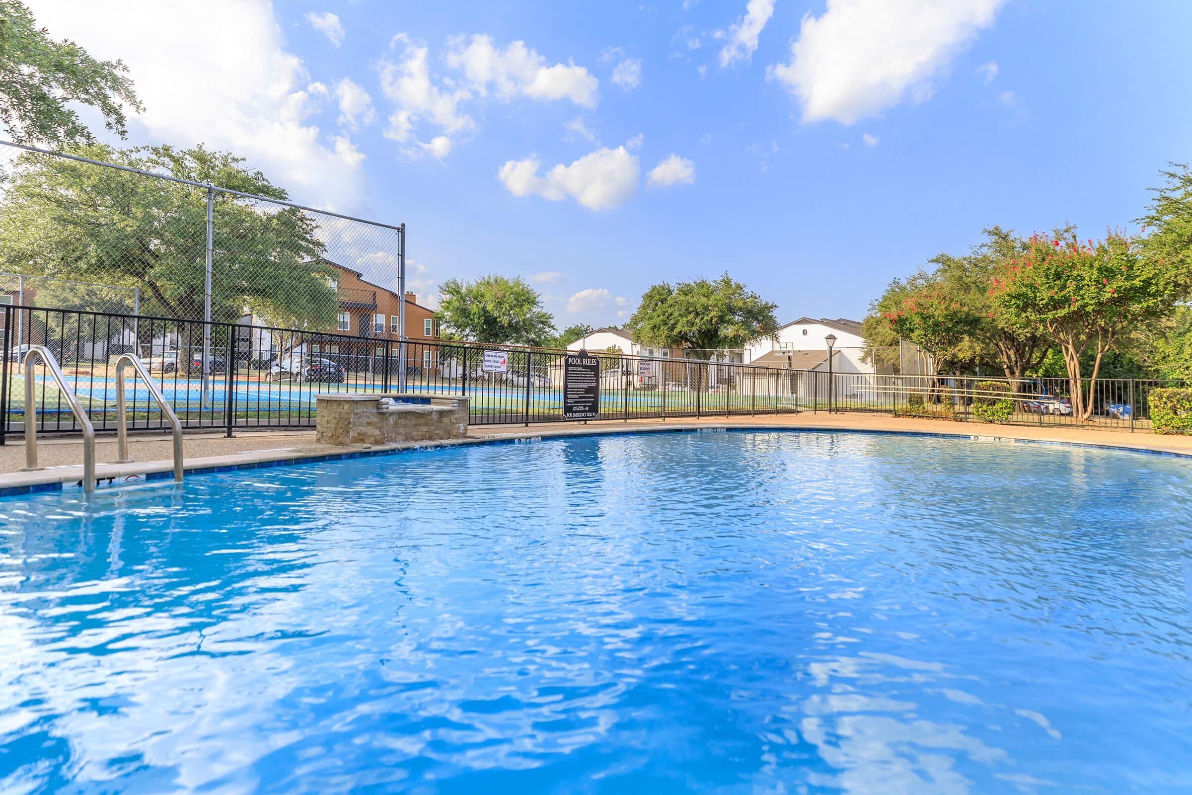 A clear blue swimming pool surrounded by a black metal fence, with trees and a few residential buildings in the background. The sky is partly cloudy, adding a serene atmosphere to the scene.
