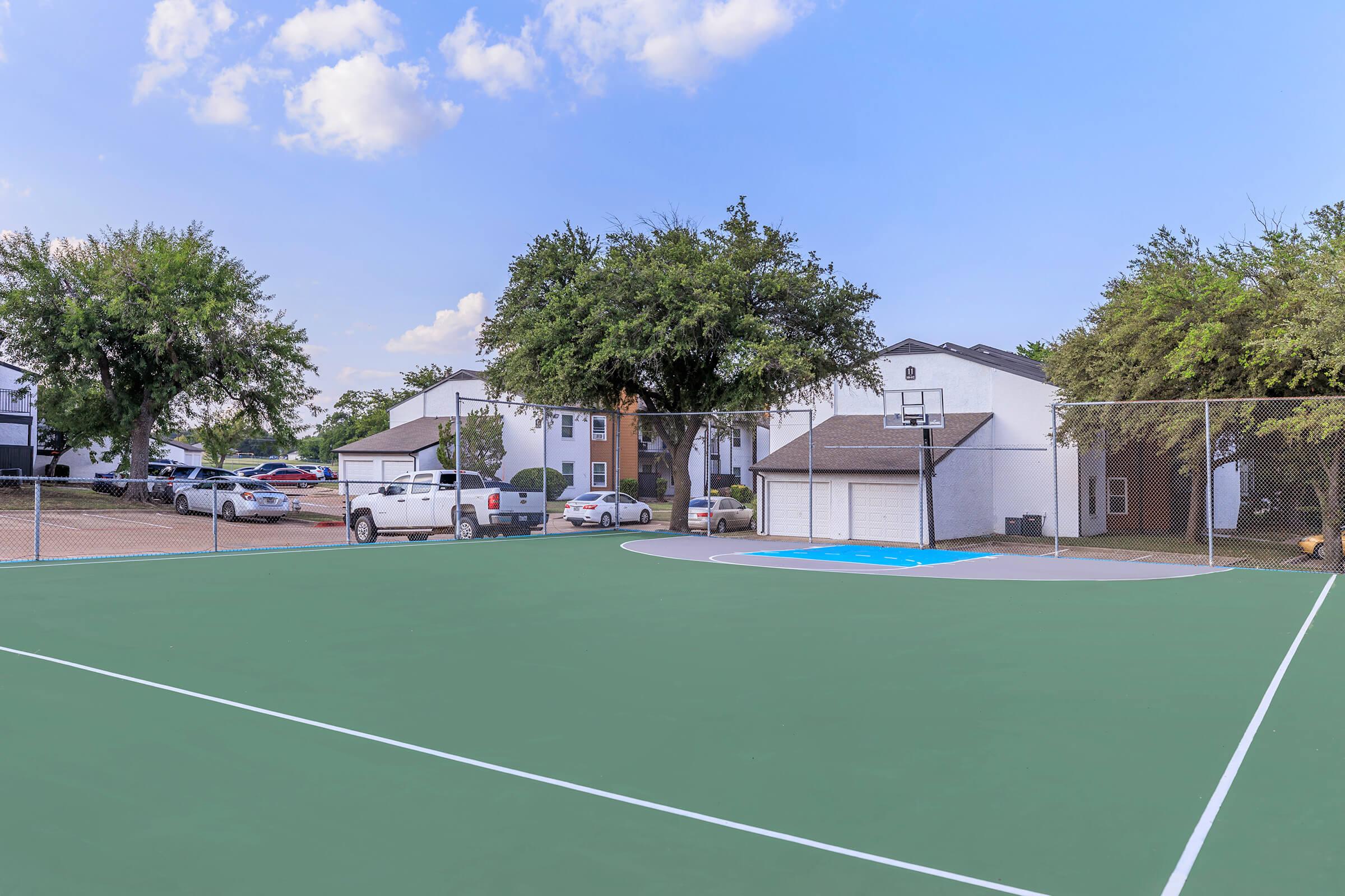 A green basketball court with a hoop in the background, surrounded by trees and residential buildings. A parking area is visible with several vehicles. The scene is bright and sunny with scattered clouds in the sky.