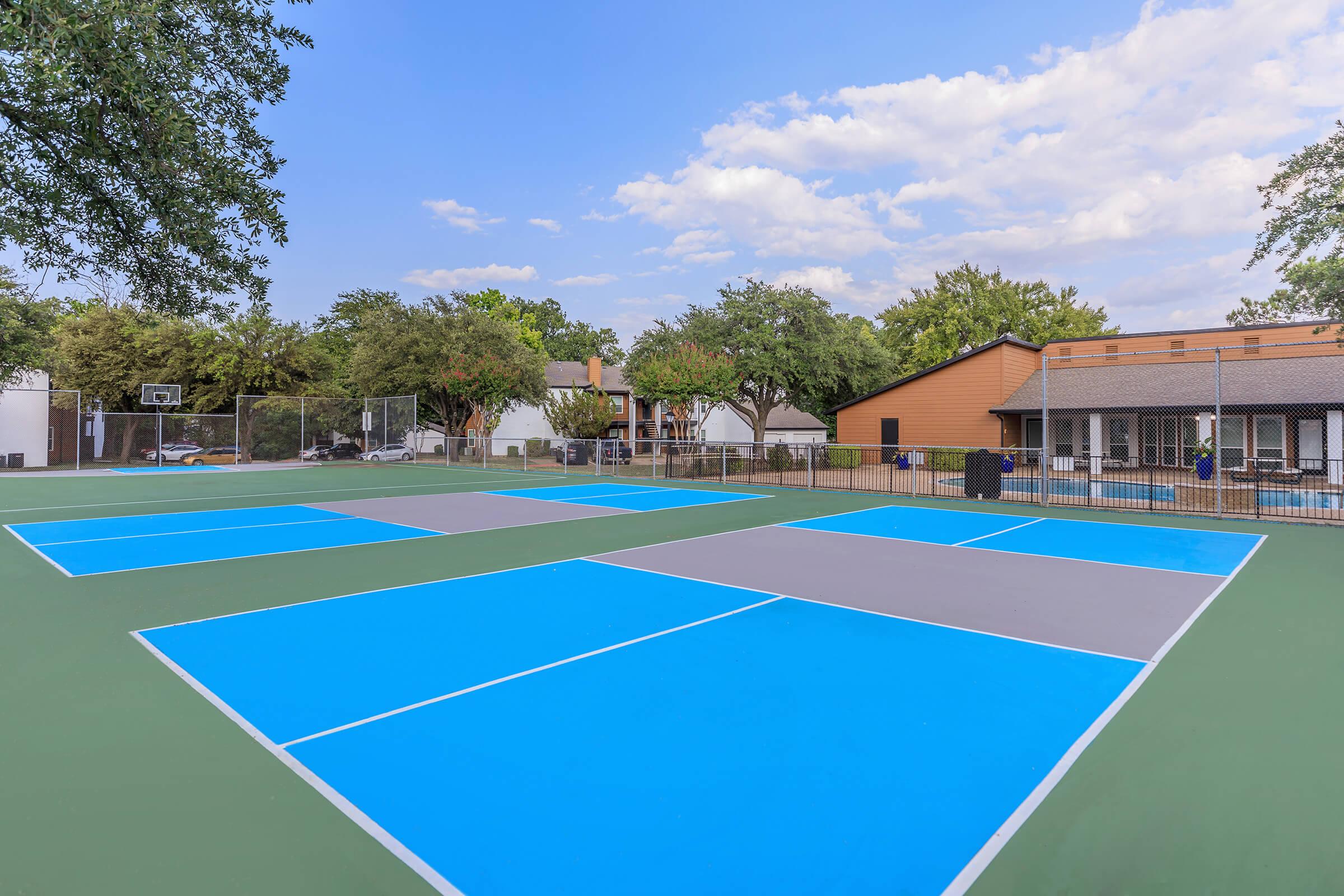 A multi-court sports area featuring several blue and gray tennis courts, surrounded by trees and a residential building. In the background, there's a basketball hoop and some shaded seating areas. The sky is partly cloudy, contributing to a bright and inviting atmosphere.