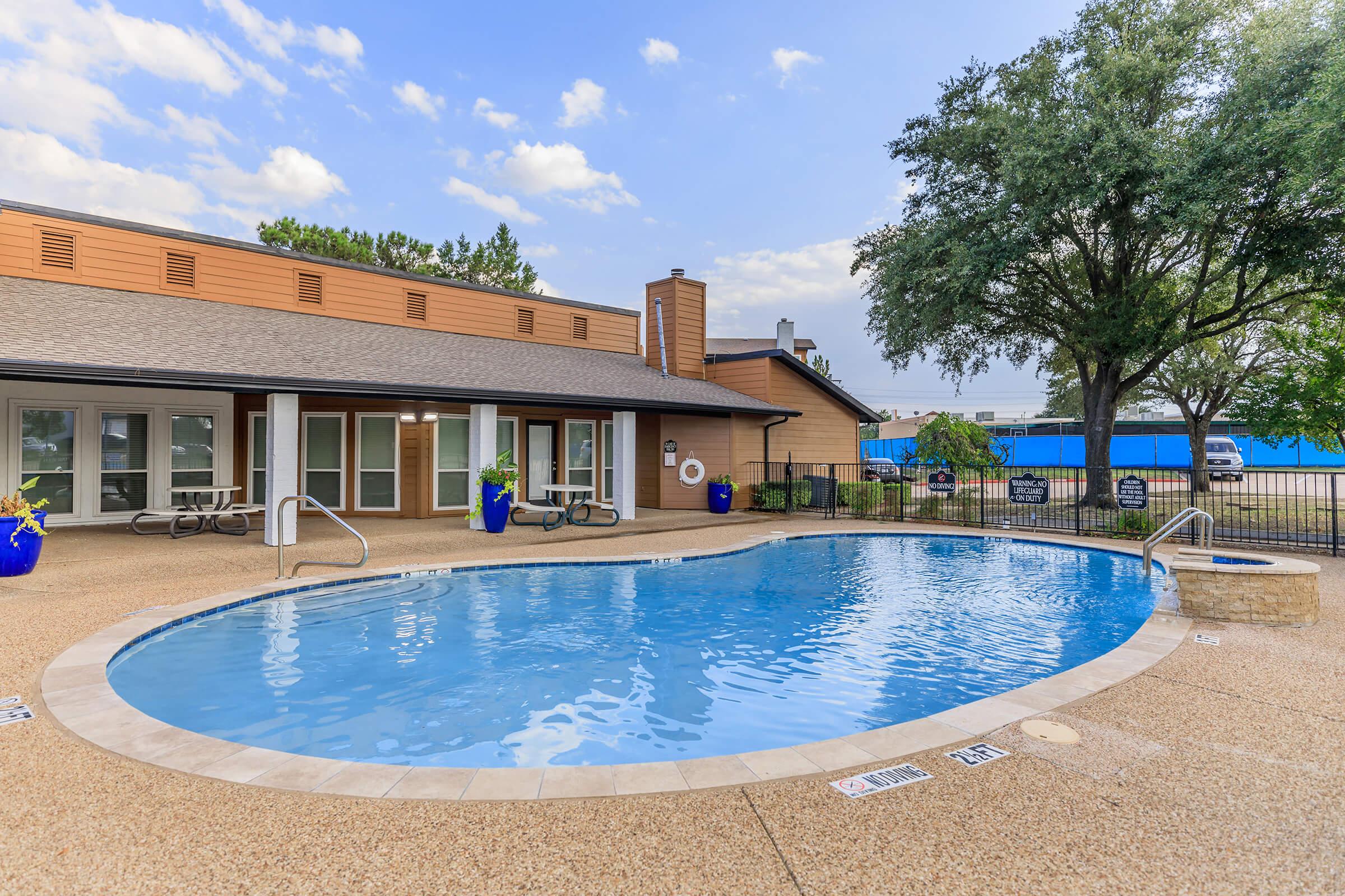 A clear swimming pool surrounded by a deck, with a light-colored building featuring large windows in the background. Lush greenery and potted plants enhance the outdoor setting. The sky is partly cloudy, and there are lounge chairs near the pool area.