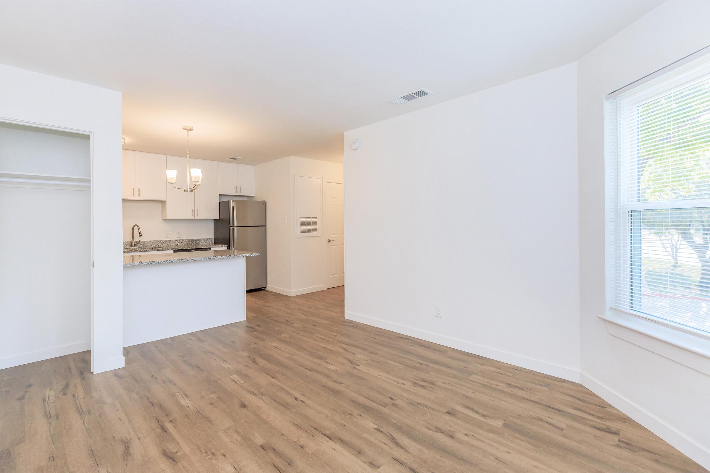 Bright and modern living space featuring light-colored walls, sleek wood flooring, and a kitchen area with granite countertops. There is a closet on the left, and large windows provide natural light. The design is open and airy, ideal for a cozy home environment.