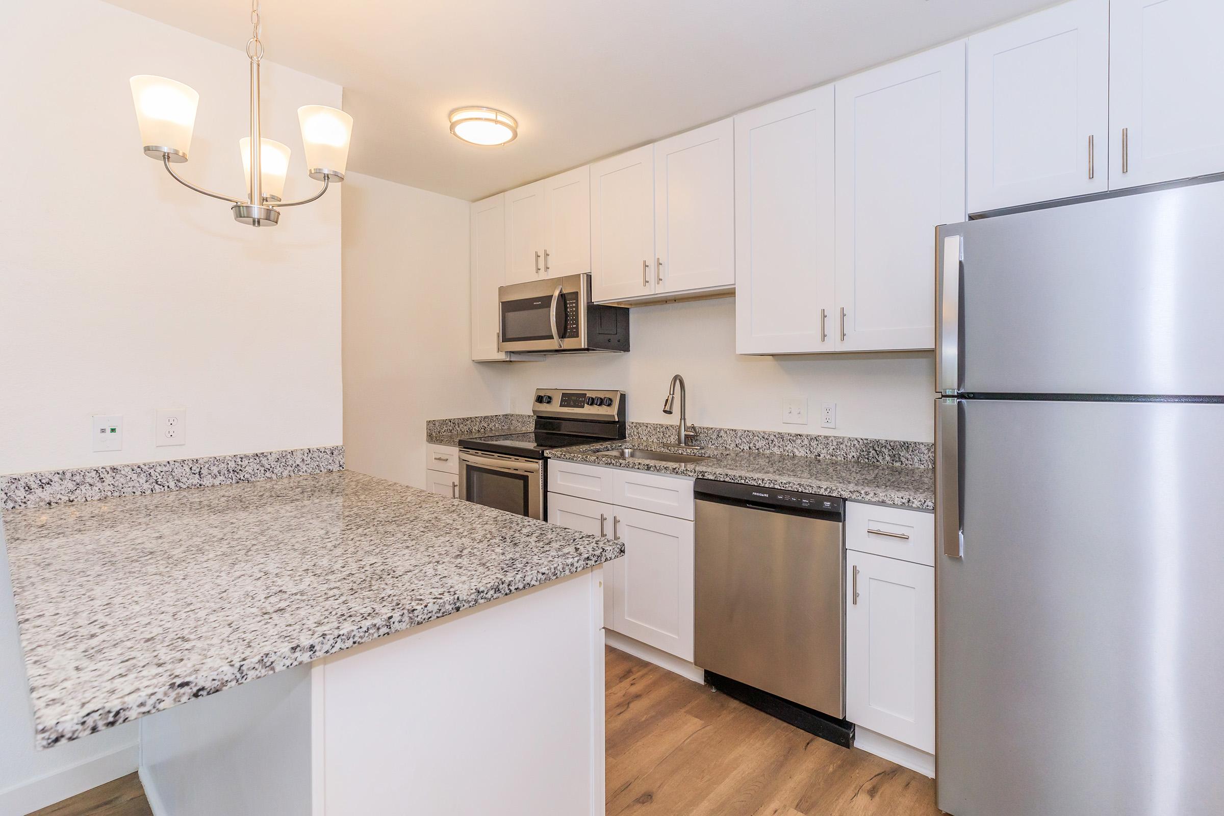 Modern kitchen featuring white cabinets, stainless steel appliances, and a granite countertop. Includes a refrigerator, oven, microwave, and dishwasher. A light fixture hangs above the island, complementing the clean and contemporary design. The flooring is wooden, adding warmth to the space.