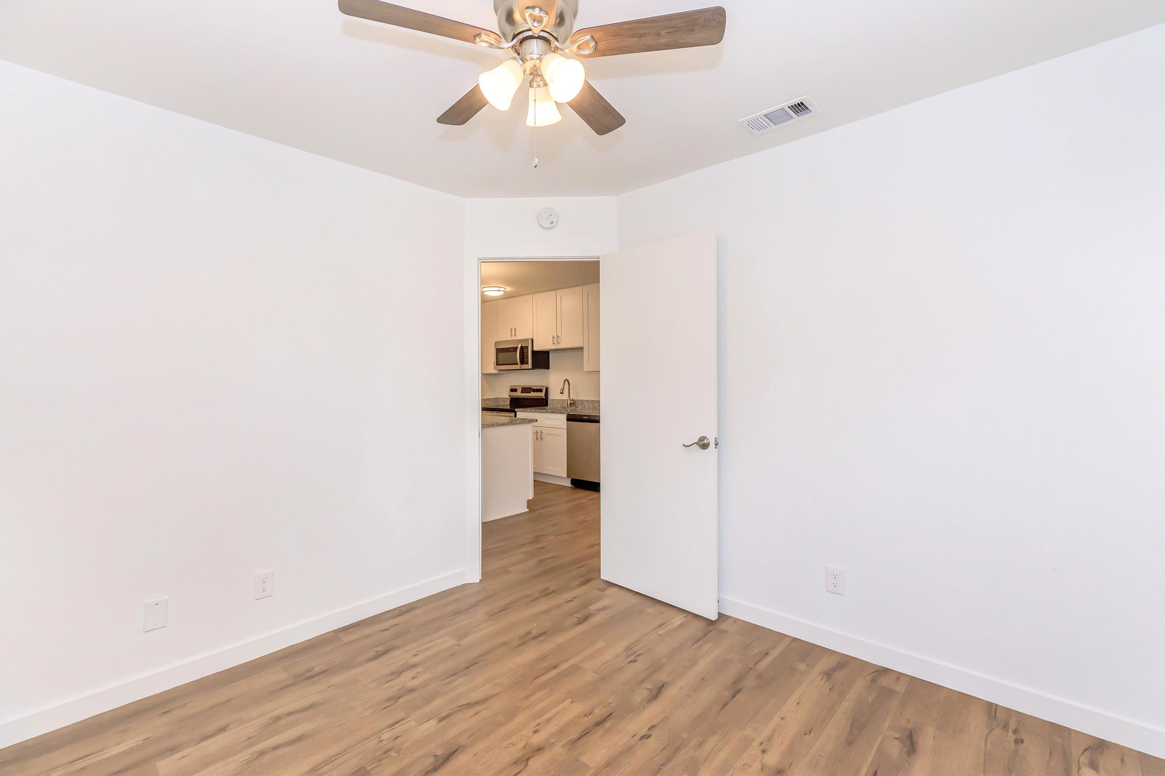 A view of an empty room featuring light-colored walls and a wooden floor. A ceiling fan with wooden blades is installed, and an open doorway leads to a kitchen area with visible appliances. The space is well-lit and has a modern appearance, suggesting it's a fresh or newly renovated environment.