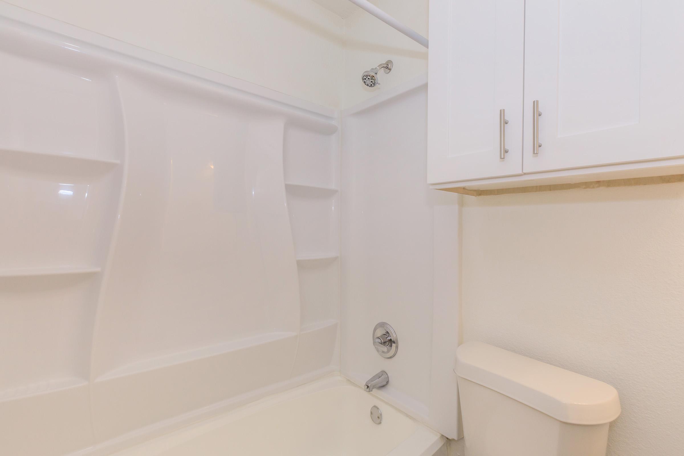 A clean, modern bathroom featuring a white bathtub and shower unit with built-in shelving. The walls are painted in a light color, and there is a white toilet beside the tub. The overall design is minimalistic and bright, enhancing the spacious feel of the room.