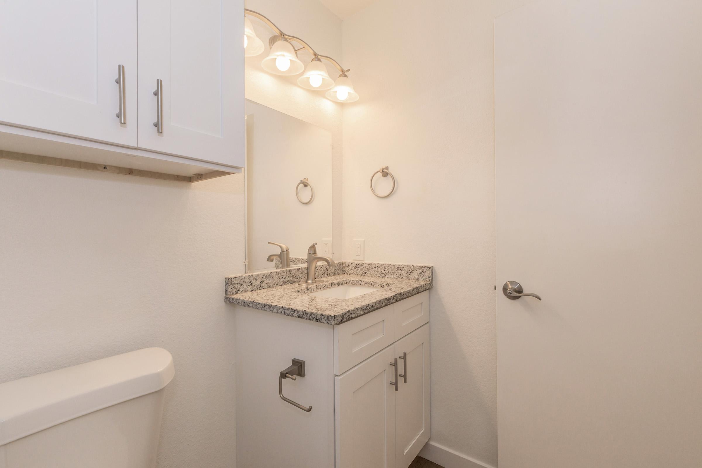 A modern bathroom interior featuring a clean, white design. The space includes a sleek sink with a granite countertop, a mounted mirror with overhead lighting, a towel ring, and a white cabinetry. A closed toilet is visible on the left, and a light-colored door is partially seen on the right.