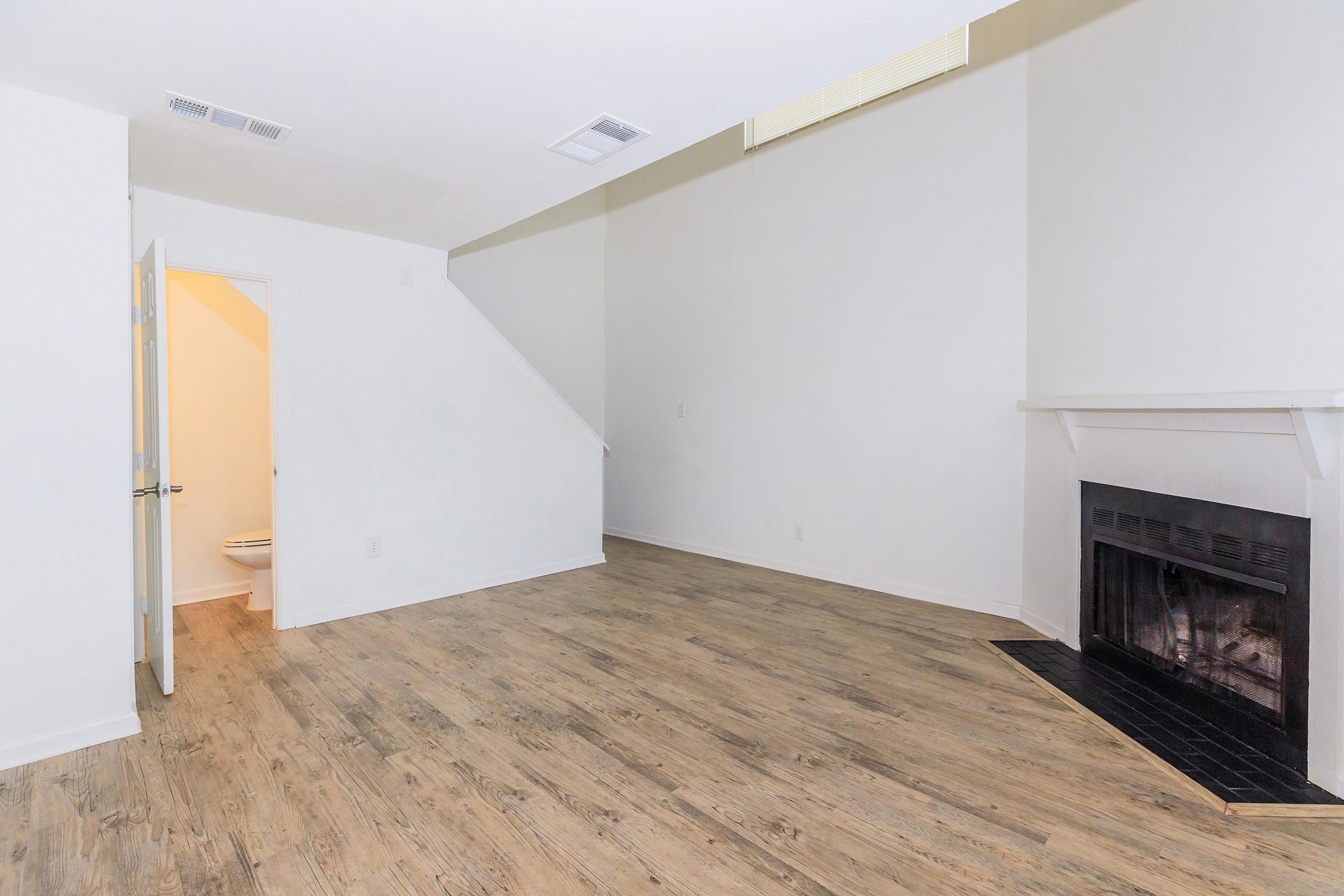 A spacious, empty living room featuring light wooden flooring, a fireplace with a black surround, and a white wall. A doorway is visible leading to a bathroom, and there is a staircase in the background. Natural light enters through a window near the ceiling.