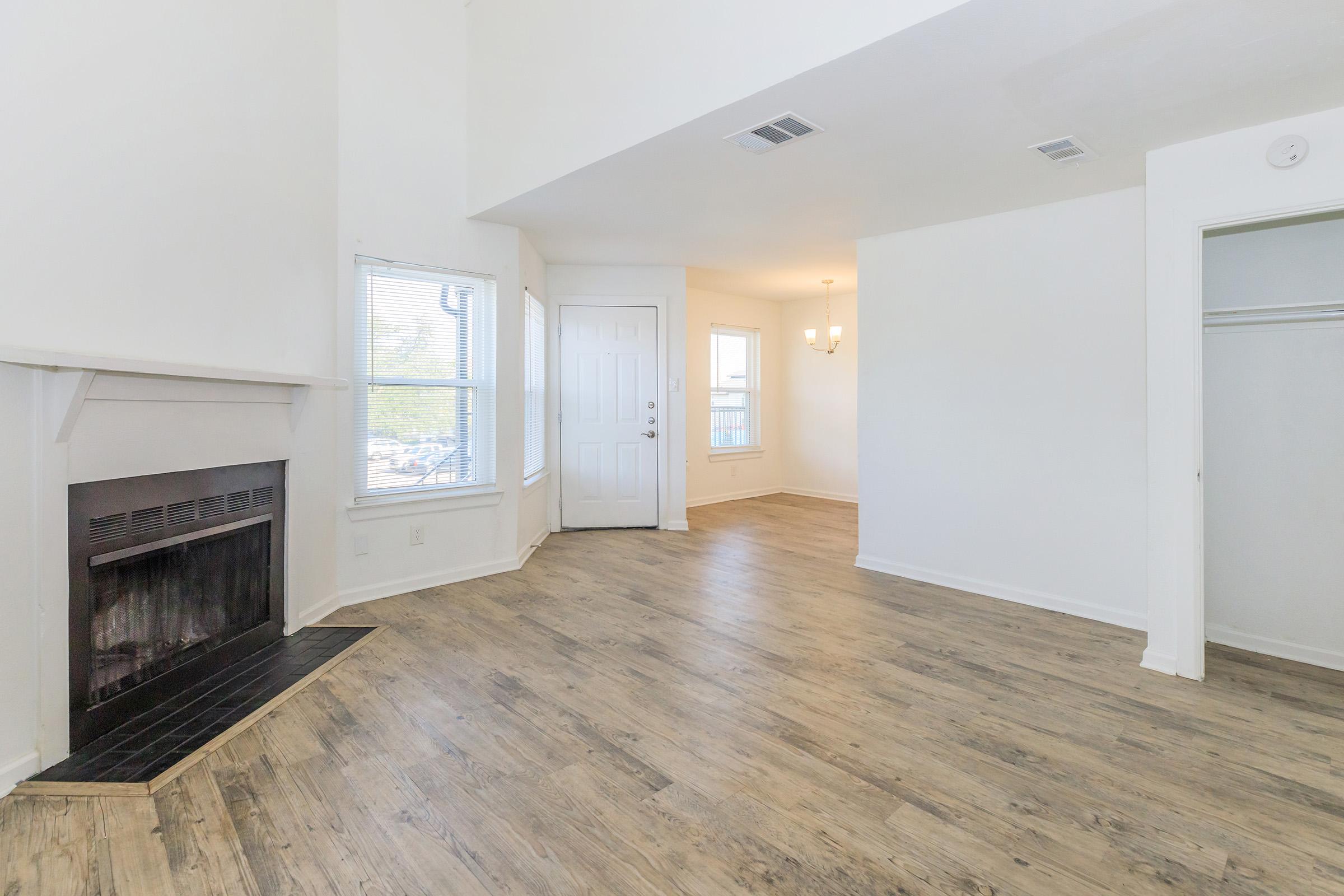 Spacious interior of a modern living area featuring a fireplace, large windows, and light-colored walls. The room includes a front door, a closet, and a bright chandelier hanging from the ceiling, with wooden flooring throughout.