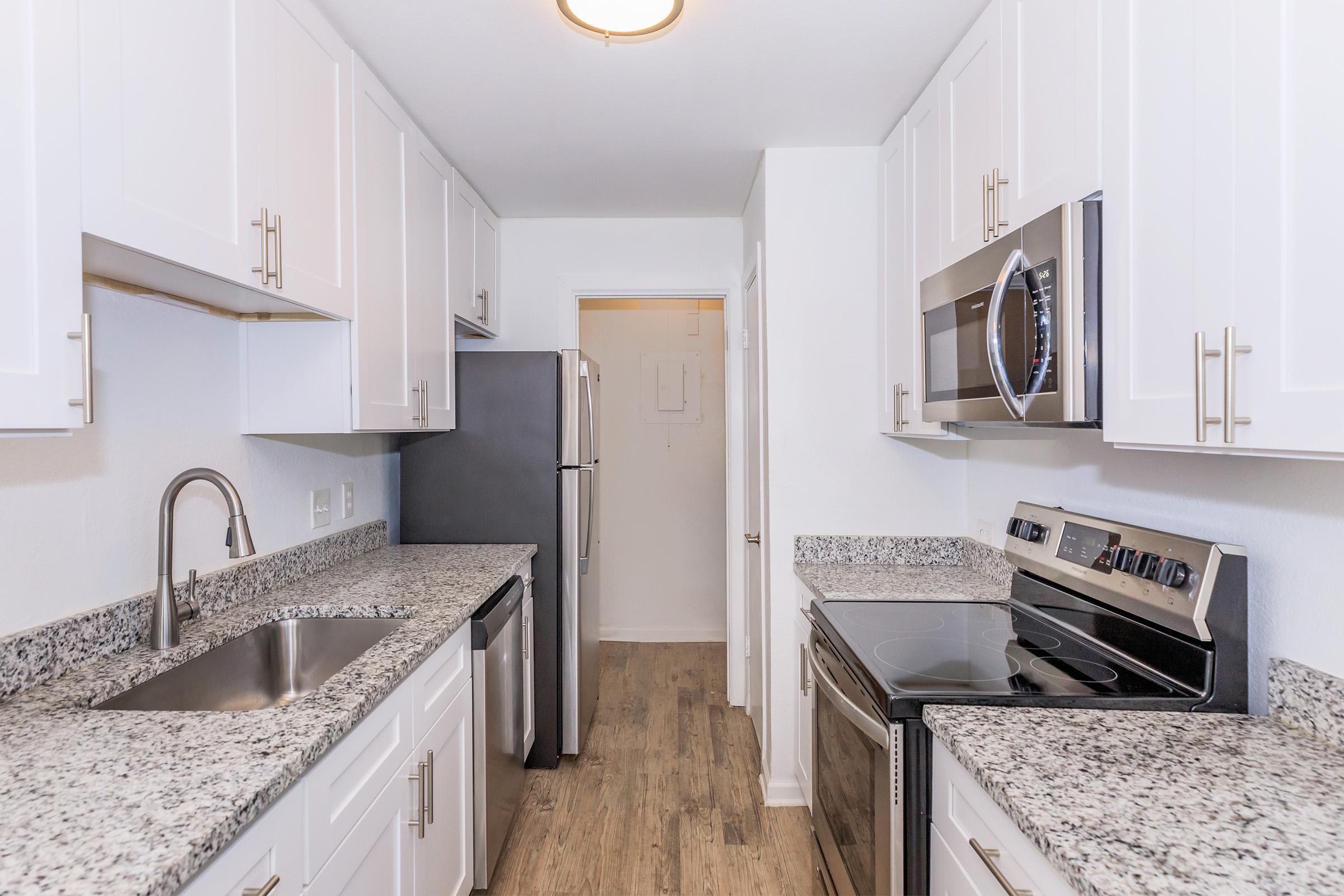 A modern kitchen featuring white cabinetry, stainless steel appliances including a refrigerator, microwave, and oven, and a granite countertop. The kitchen has a spacious layout with a sink and is well-lit, showcasing a clean and contemporary aesthetic.