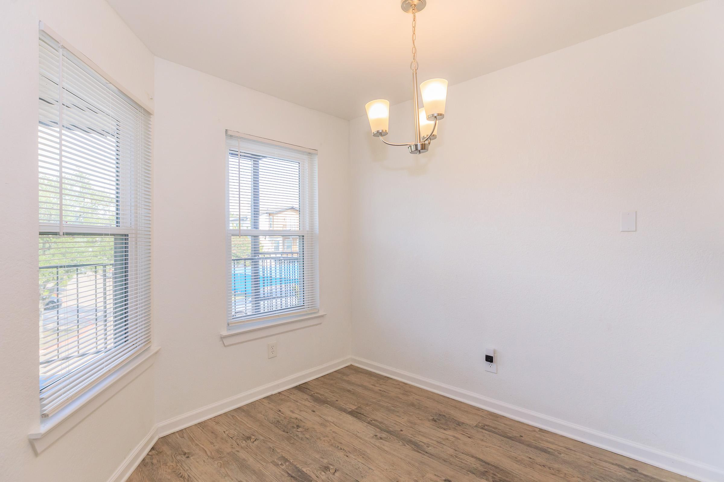 A bright, empty room featuring two windows with blinds and a ceiling light fixture. The walls are painted white, and the flooring is light-colored wood. The space is uncluttered and offers natural light, creating a clean and airy atmosphere.