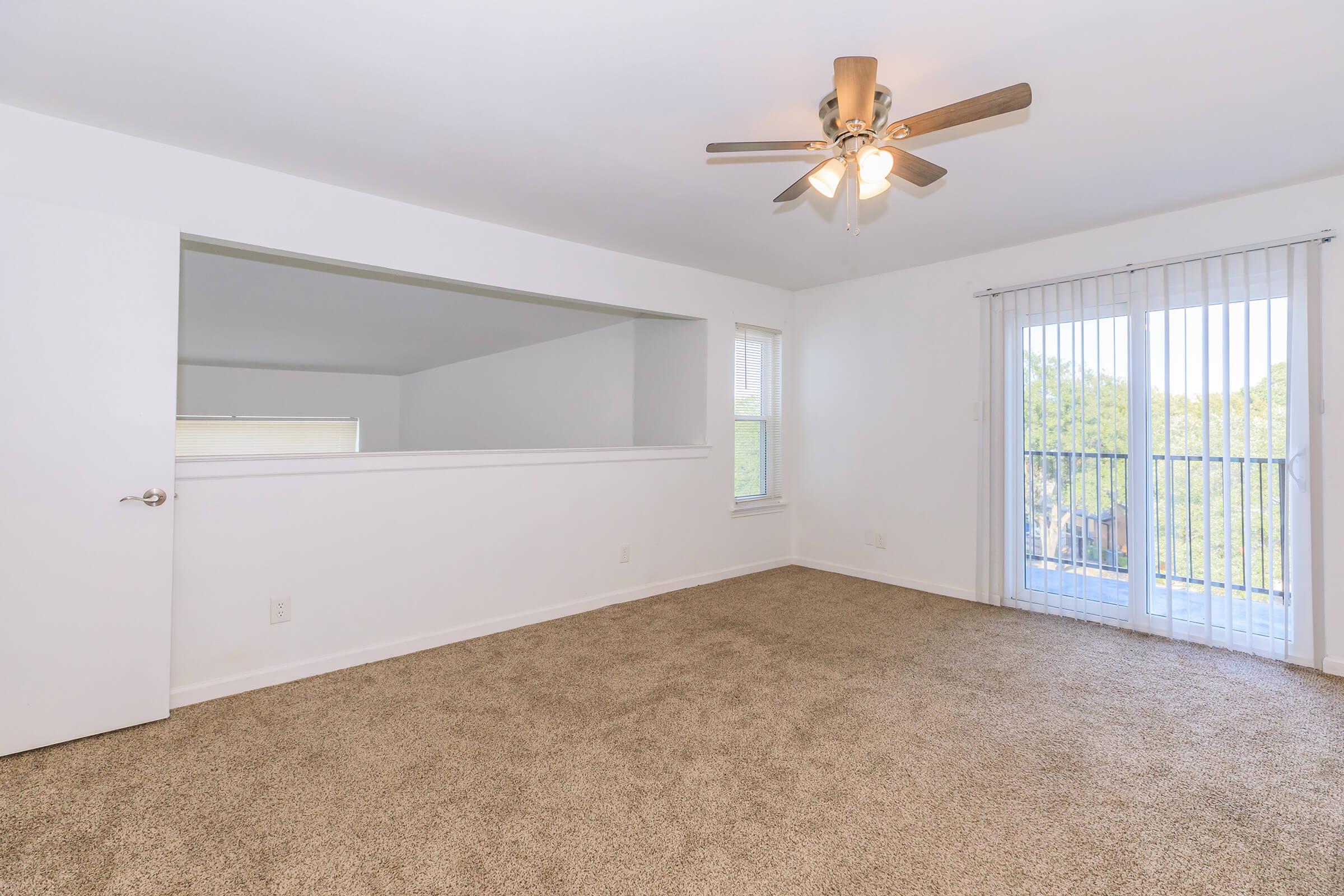 Bright and airy living room featuring beige carpet, a ceiling fan, and natural light from large sliding glass doors. A partition divides the room, creating an open feel. Walls are painted white, and the space is ready for personalized decor.