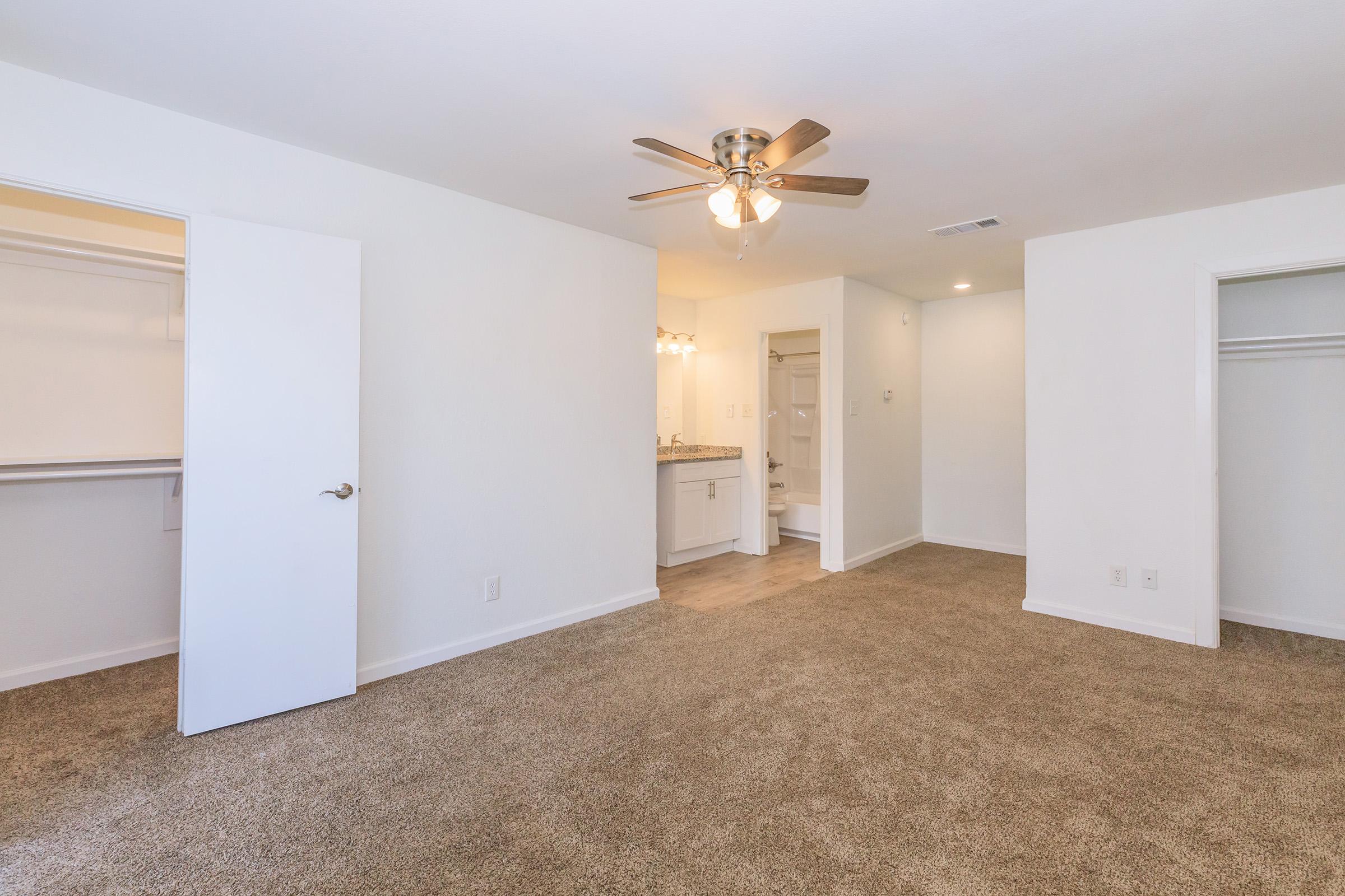 A spacious interior room with beige carpet, featuring two open doors leading to closets and a modern bathroom area with a vanity. The room has a ceiling fan and neutral-colored walls, creating a bright and inviting atmosphere.