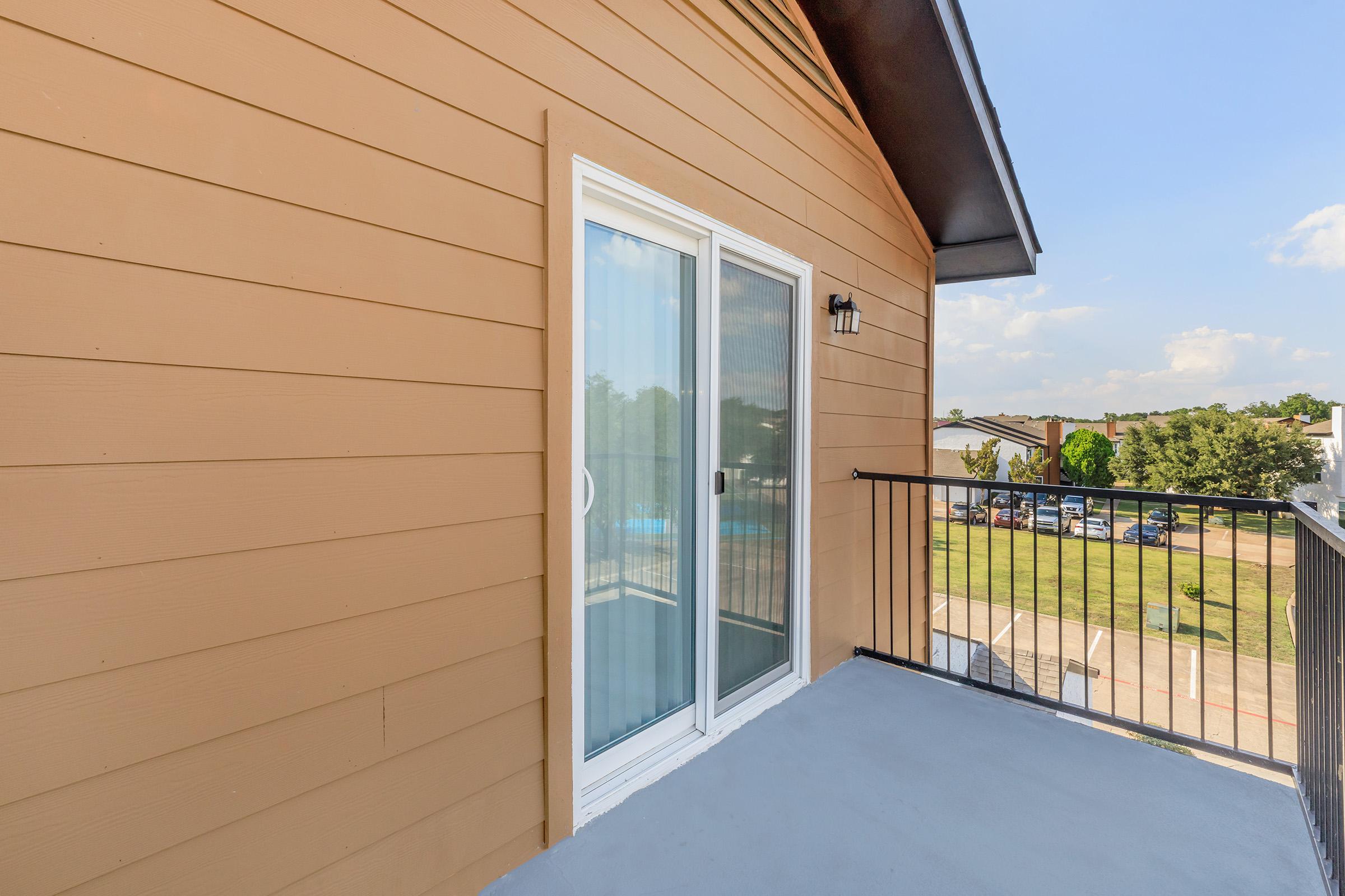 A residential balcony with a sliding glass door providing access to an outdoor space. The wall features brown siding, and the balcony has a metal railing. In the background, there are trees and parked cars visible. The sky is partly cloudy, suggesting a pleasant day.