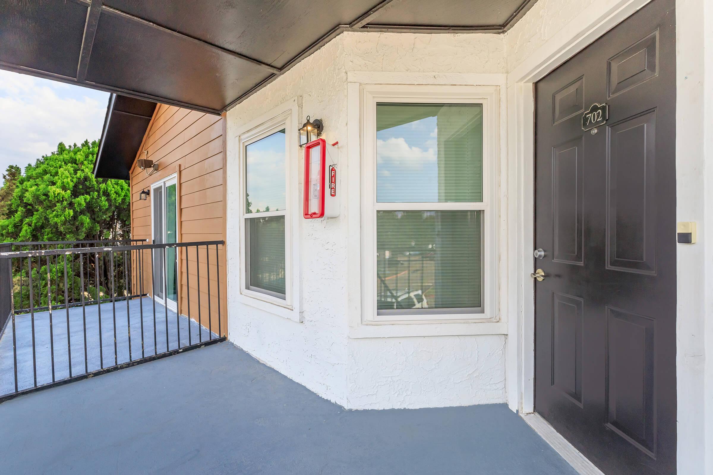 Exterior view of an apartment entrance featuring a black door with the number 702, two windows beside it, and a small balcony area. The building has a combination of light-colored stucco and wood accents, with greenery visible in the background. The porch has a railing and a fire extinguisher mounted on the wall.