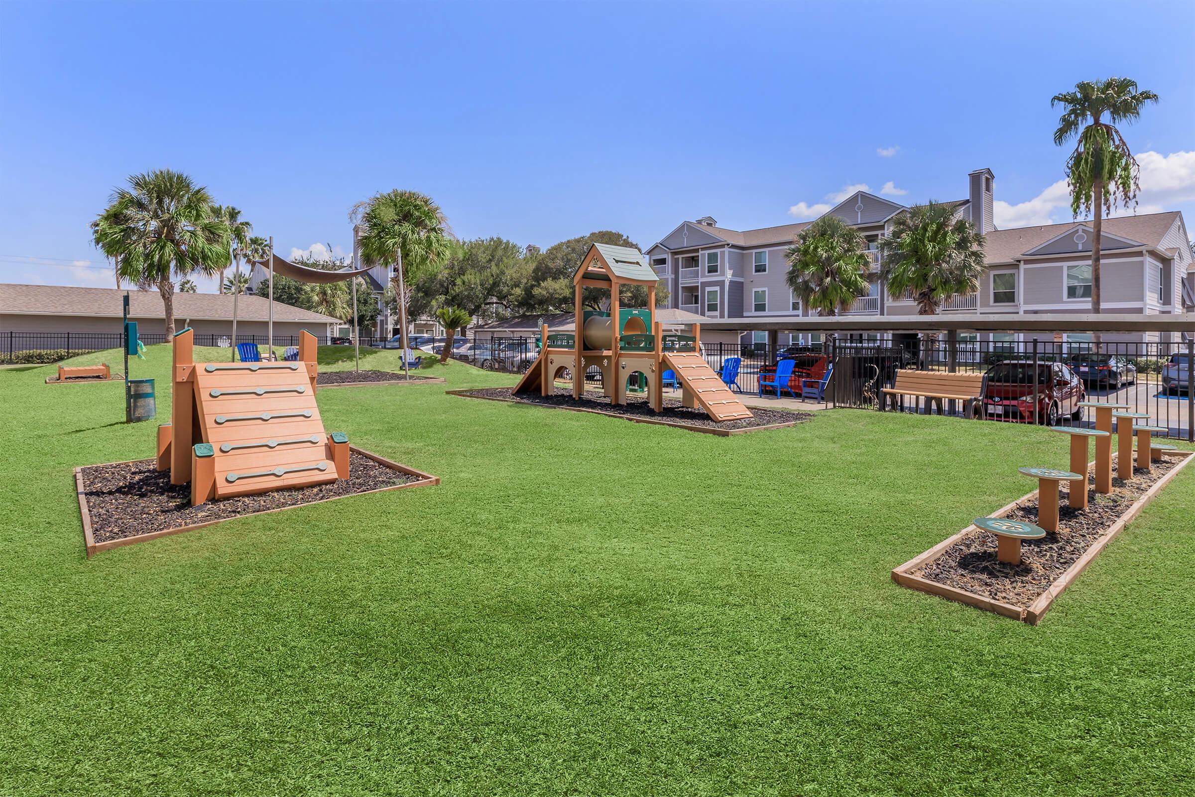 A bright playground with green artificial grass, featuring a climbing structure, slides, and play equipment. In the background, there are palm trees and a multifamily residential building. The area is fenced, creating a safe space for children to play and explore.