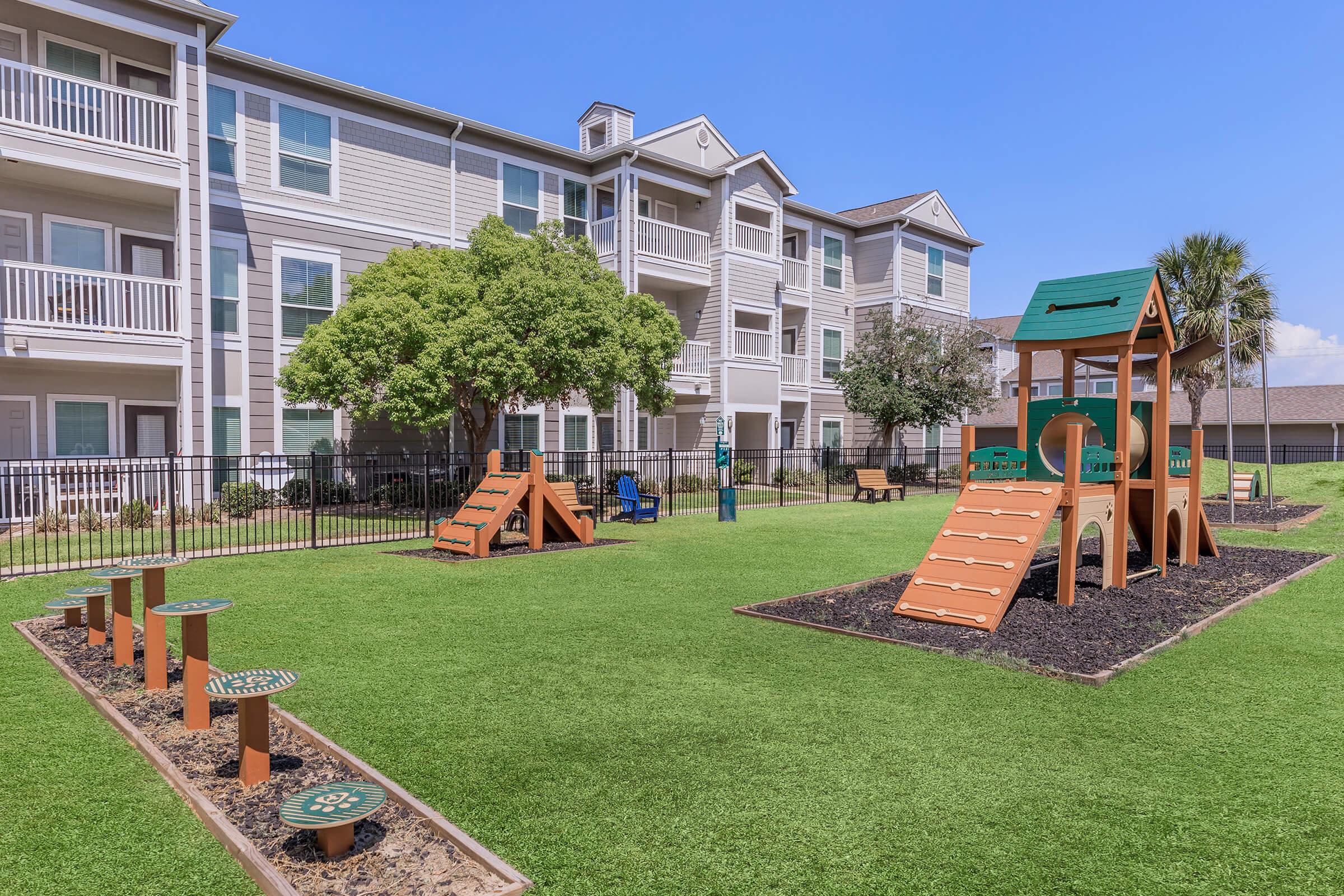 Playground area featuring a grassy lawn with two small slides and climbing structures, surrounded by a fence. In the background, there are several apartment buildings with balconies. The sky is clear and blue, providing a sunny, inviting atmosphere for outdoor activities.