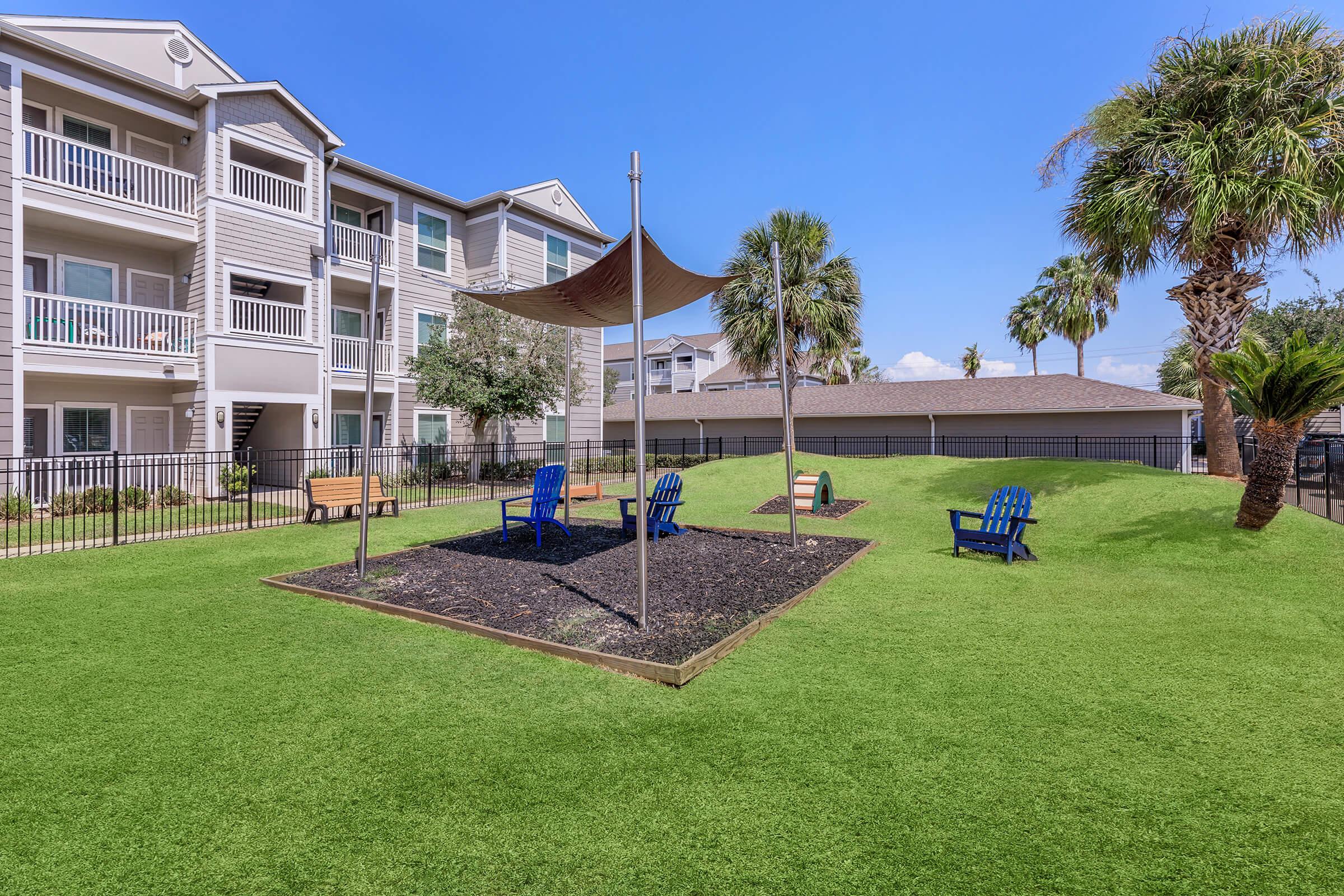 A sunny outdoor area featuring a playground with blue chairs, a shaded structure, and a green lawn. Surrounding the playground are palm trees and residential buildings, creating a pleasant atmosphere for relaxation and play.