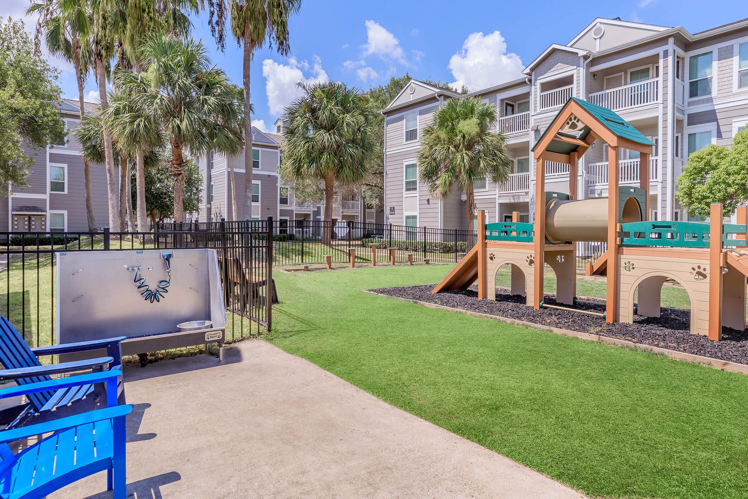 A colorful playground set within a landscaped area featuring artificial grass, surrounded by palm trees and apartment buildings. Blue seating is visible in the foreground, and a fenced area encloses the playground, creating a family-friendly atmosphere.