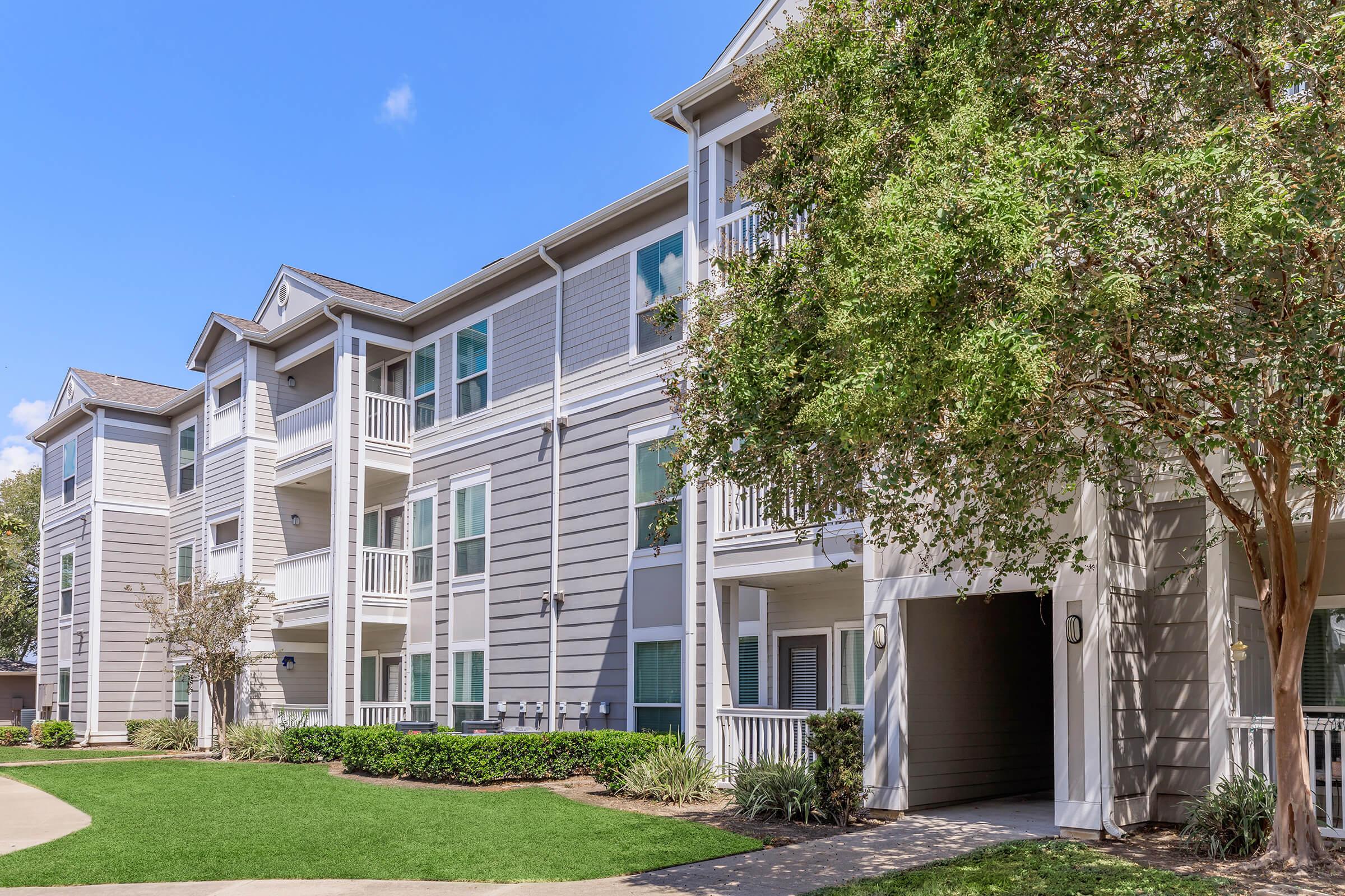 A view of a modern multi-story apartment building with a light gray exterior, featuring balconies and landscaped grounds. There are green lawns and shrubs in front, creating a welcoming environment under a clear blue sky. The scene conveys a pleasant residential setting.