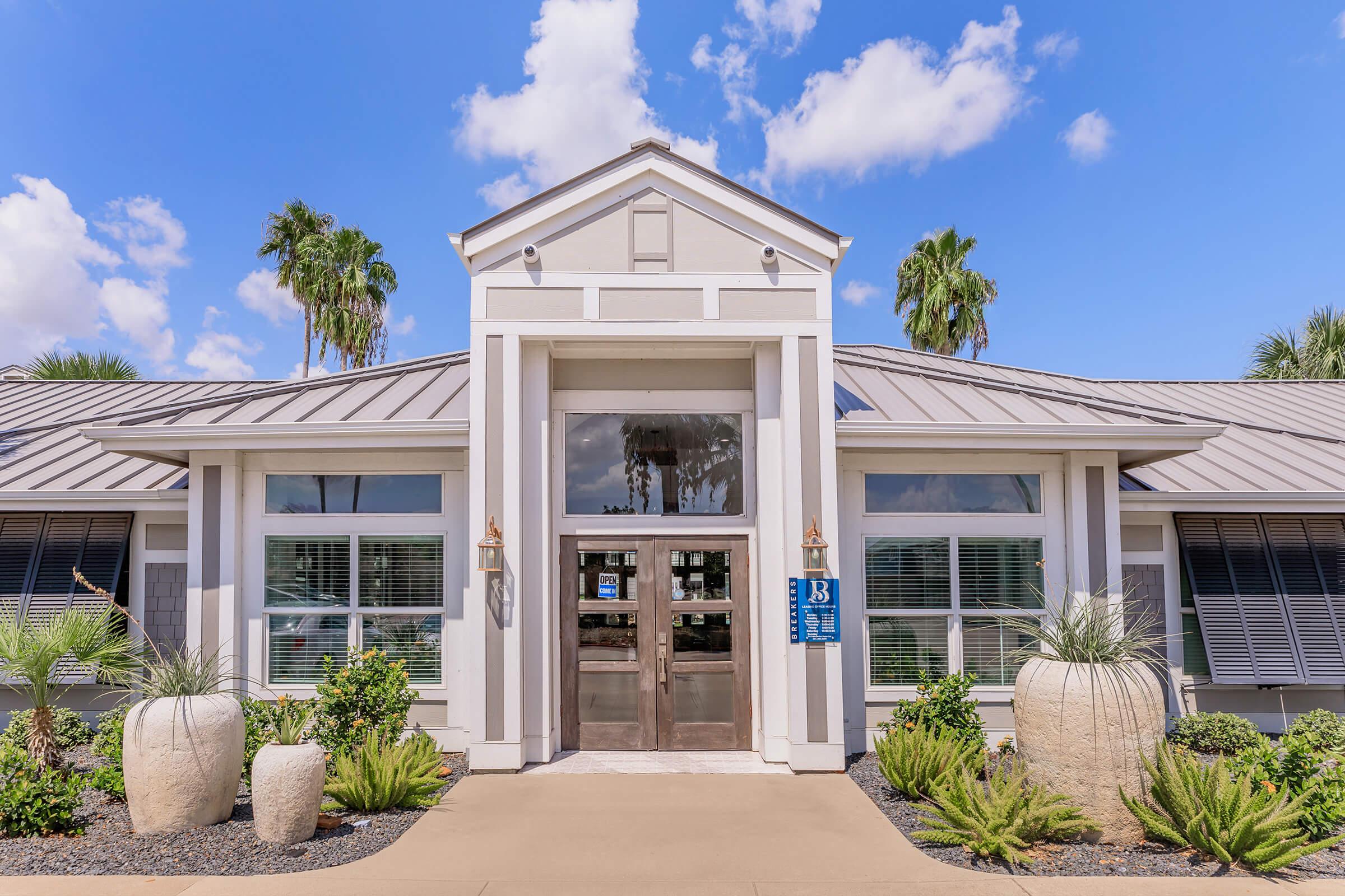 A modern building with a symmetrical design, featuring a large entrance with wooden double doors. The facade is painted in light colors, accented by large windows. Flanking the entrance are decorative planters with lush greenery. Above, a clear blue sky with fluffy clouds complements the scene. Palm trees are visible nearby.