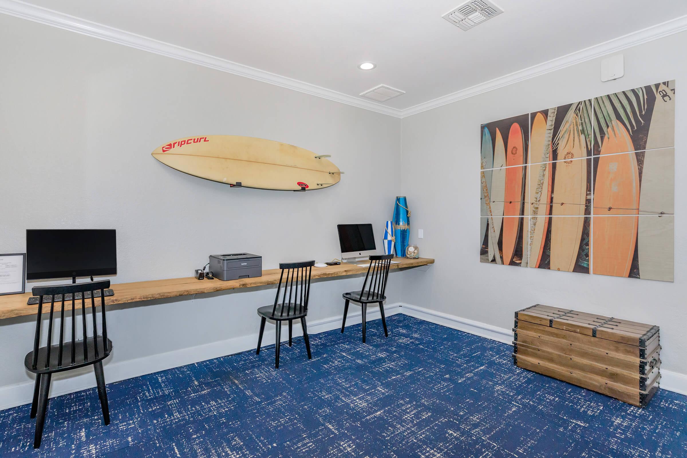 A modern workspace featuring two black chairs at a long wooden desk with two desktop computers. A surfboard hangs on the wall above the desk, and a surfboard-themed art piece is displayed on another wall. The room has blue carpet and light gray walls, creating a bright and airy atmosphere.