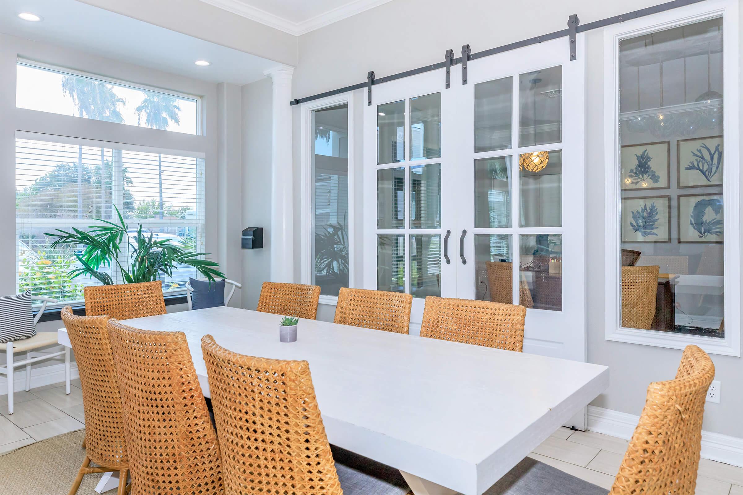 A bright, modern dining room featuring a large white table surrounded by woven chairs. Large windows let in natural light, showcasing greenery outside. Decorative wall art can be seen in the background, and sliding glass doors separate the dining area from another room.