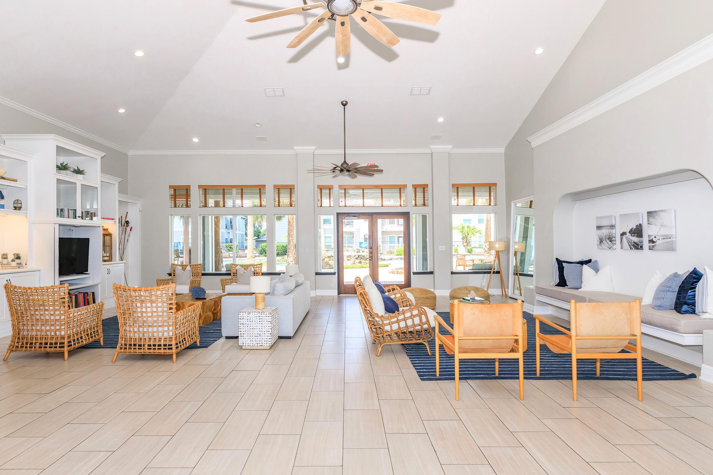 Spacious, modern living room featuring stylish wicker and cream-colored furniture. Large windows allow natural light, with a view of outdoor greenery. The floor is tiled, and decor includes light wood accents and blue textiles. A ceiling fan and light fixtures add a cozy ambiance.