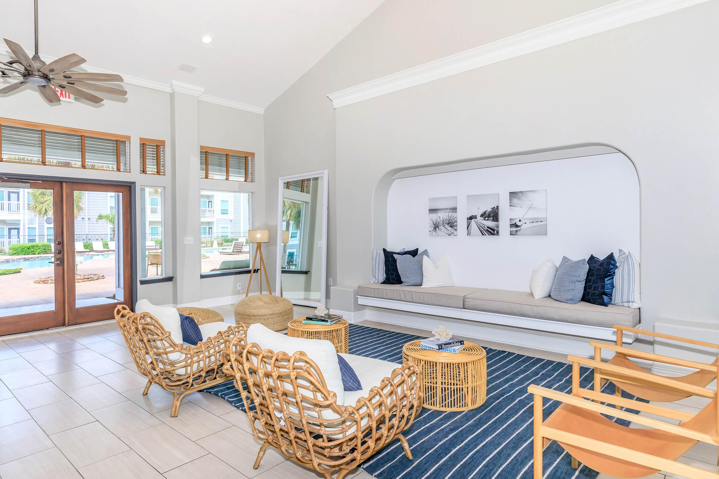 Bright and airy lounge area featuring two rattan armchairs, a beige sofa with decorative pillows, a coffee table, and a woven pouf. The walls are painted in light gray with three framed black-and-white photographs. Large windows allow natural light, and a blue and white striped rug adds a cozy touch.